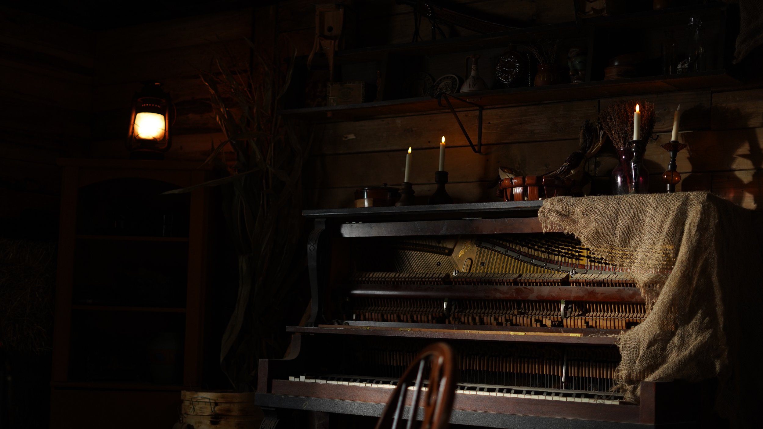 An old upright piano with its cover removed, revealing the inner strings and hammers, placed in a dimly lit rustic wooden room. On top of the piano, there are three lit candles in holders, a dark vase with dried flowers, and some decorative items. To the left, a vintage lantern emits a warm glow, casting shadows on the wooden walls.