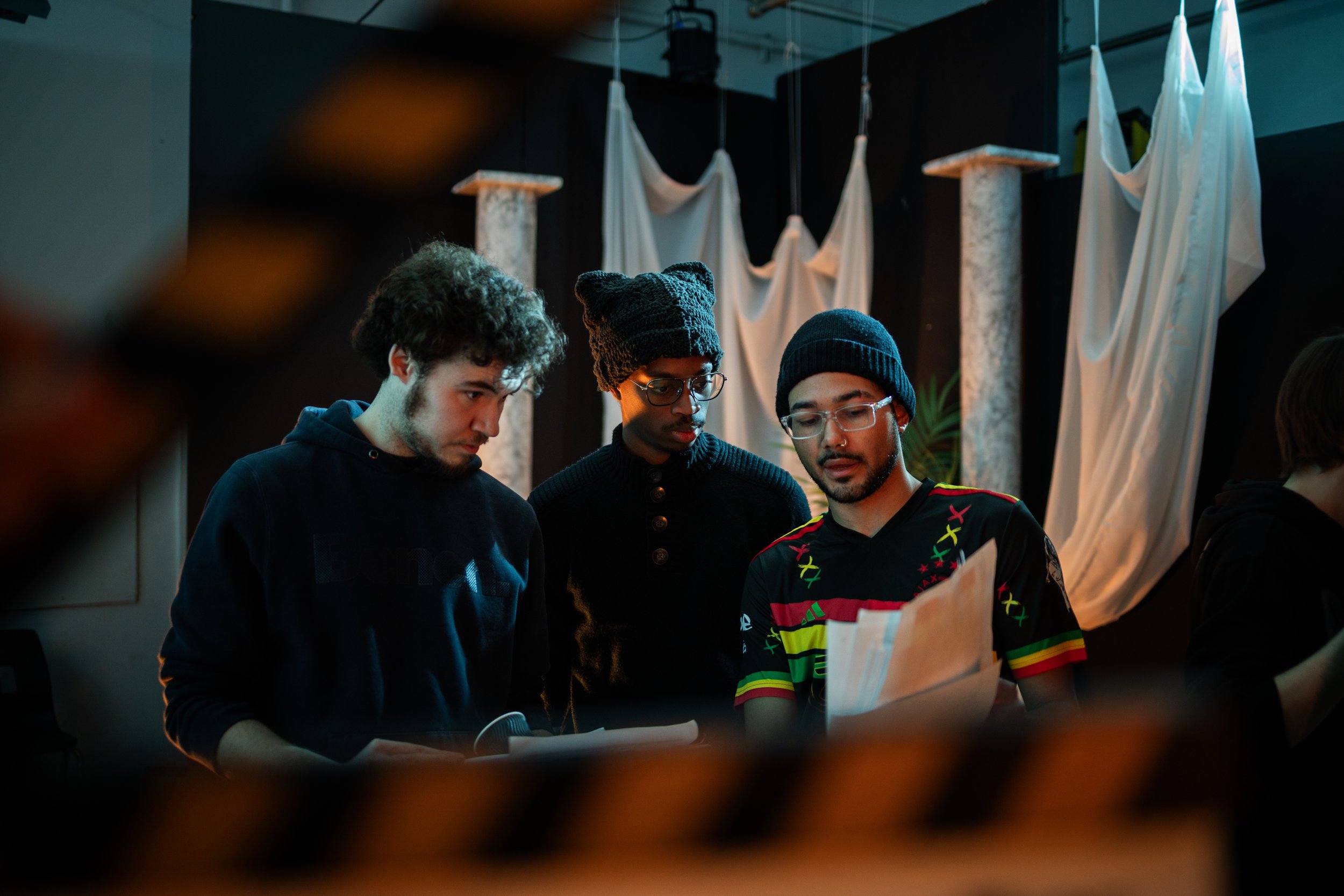Three young men inspecting papers or scripts in a dimly lit room with artistic fabric and plant decor behind them.