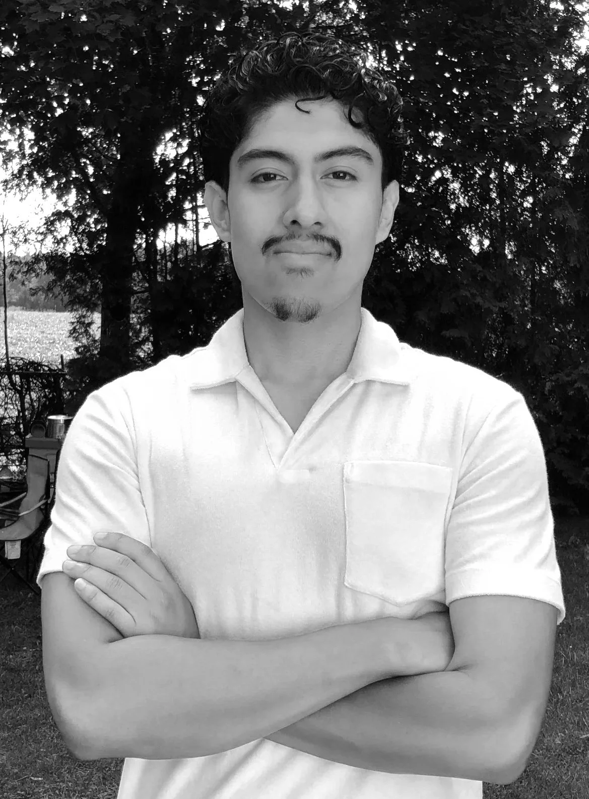A young man with curly hair, a mustache, and goatee stands outdoors with arms crossed, wearing a white polo shirt, in front of trees.