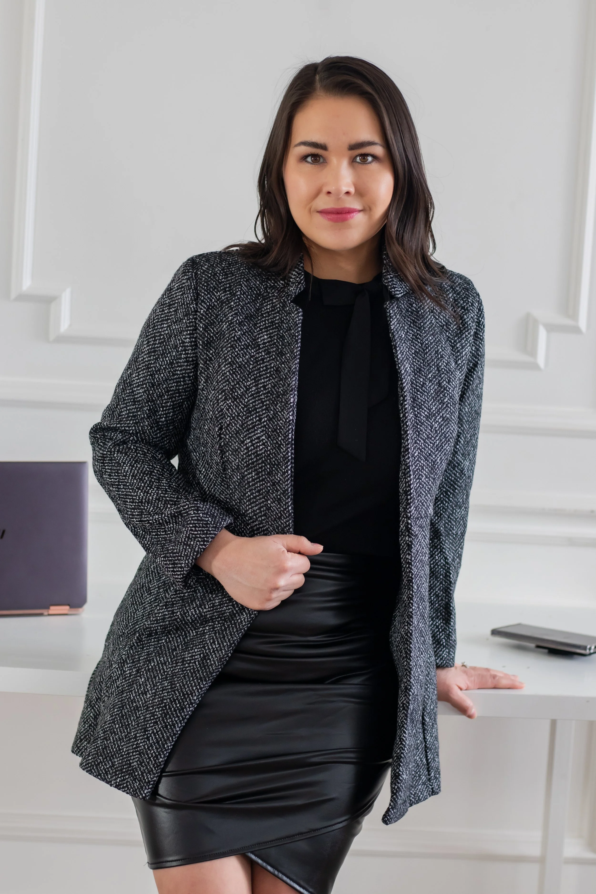 A young woman in business attire standing beside a desk with a laptop and a tablet, posing confidently in a well-lit, elegant office.