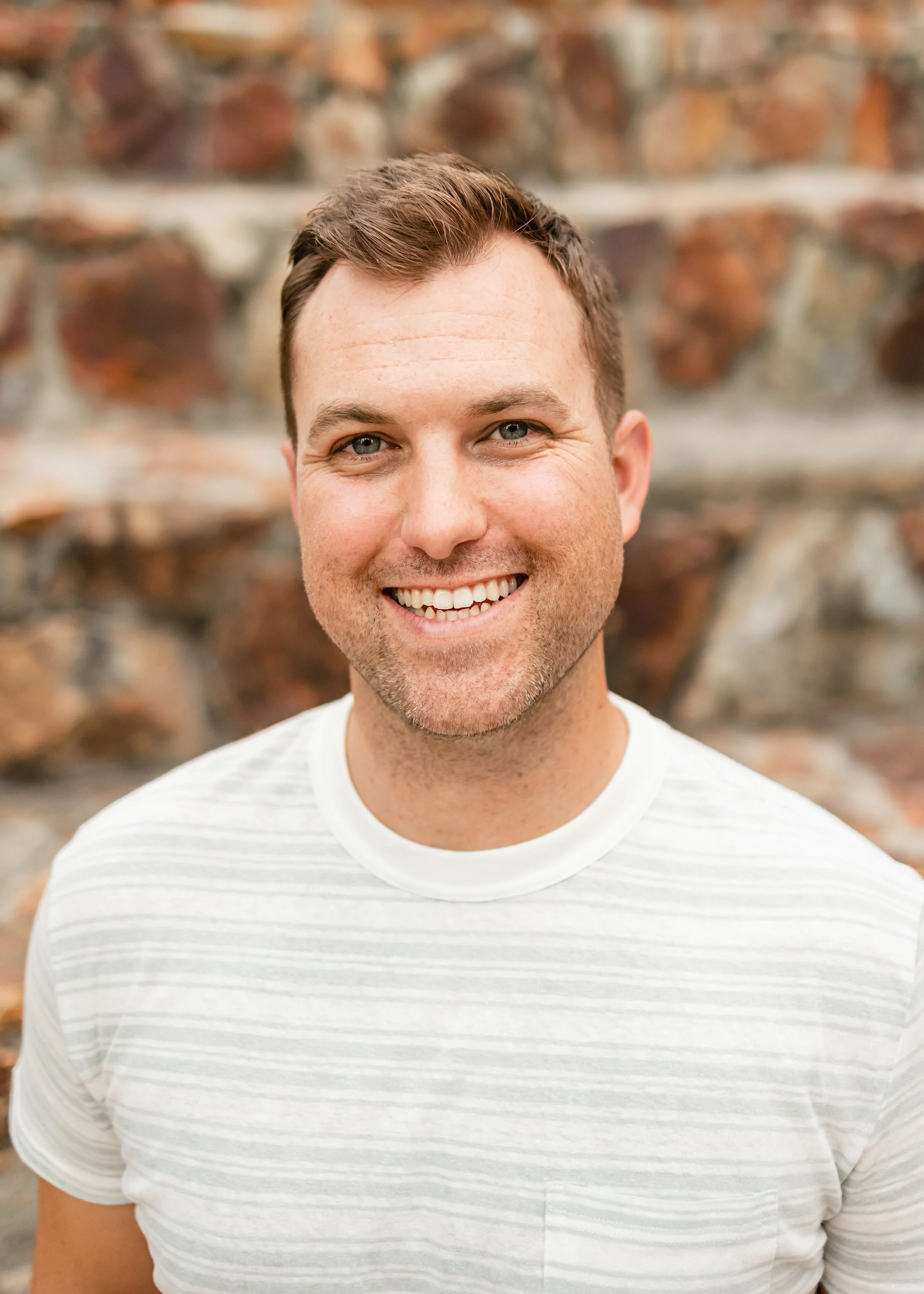 Smiling young man with short brown hair and blue eyes, wearing a white striped t-shirt, standing in front of a stone wall.