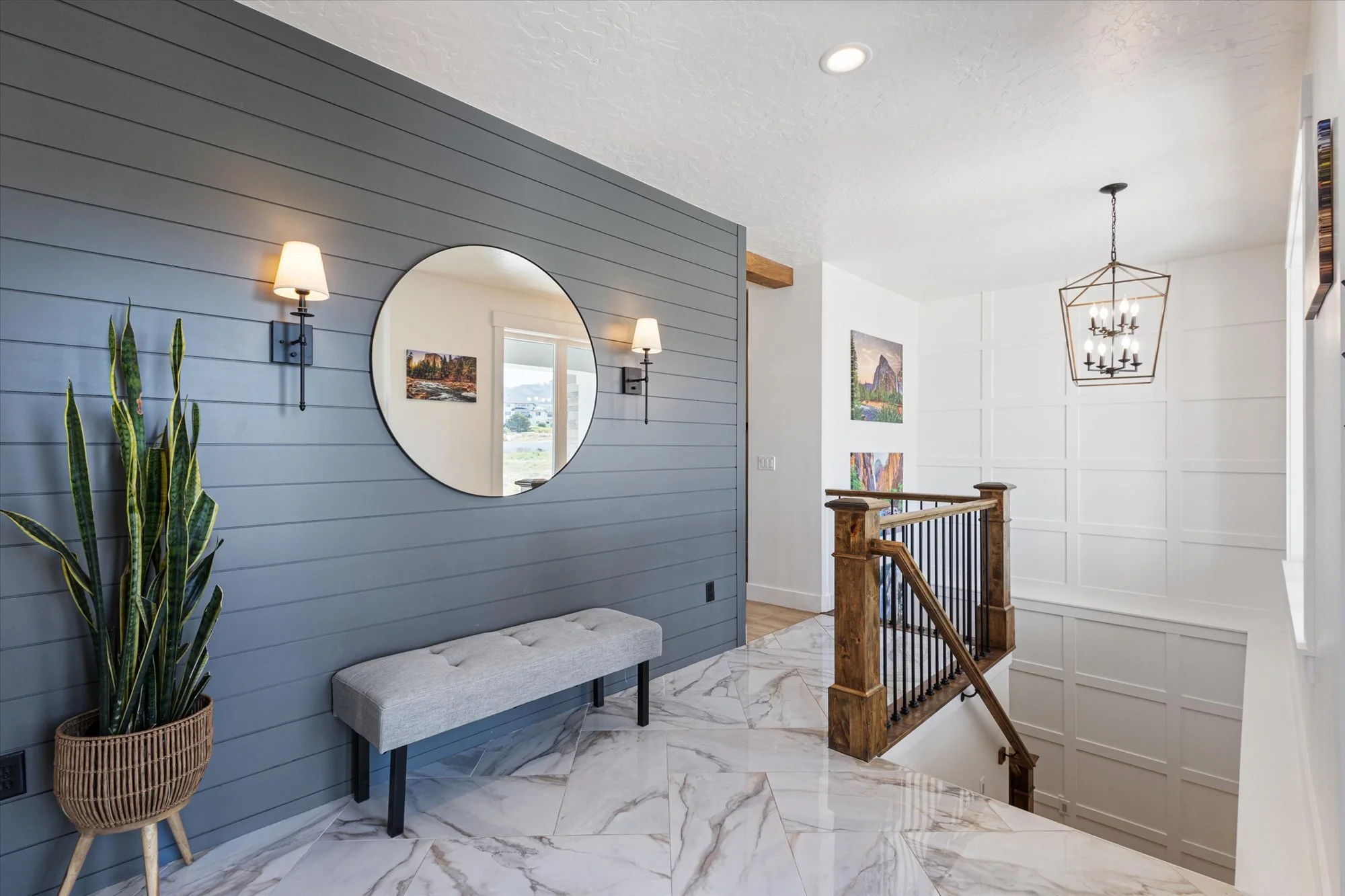 Interior view of a home entrance with a blue shiplap wall, a round mirror, wall sconces, a plant in a woven basket, a gray upholstered bench, a marble-tiled floor, a wooden staircase with black metal railing, and artwork on a white paneled wall.