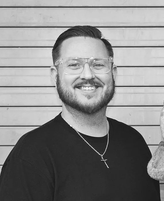 Black and white photo of a smiling man with glasses, a beard, and short hair, wearing a black shirt and a cross necklace, standing in front of a wooden wall.