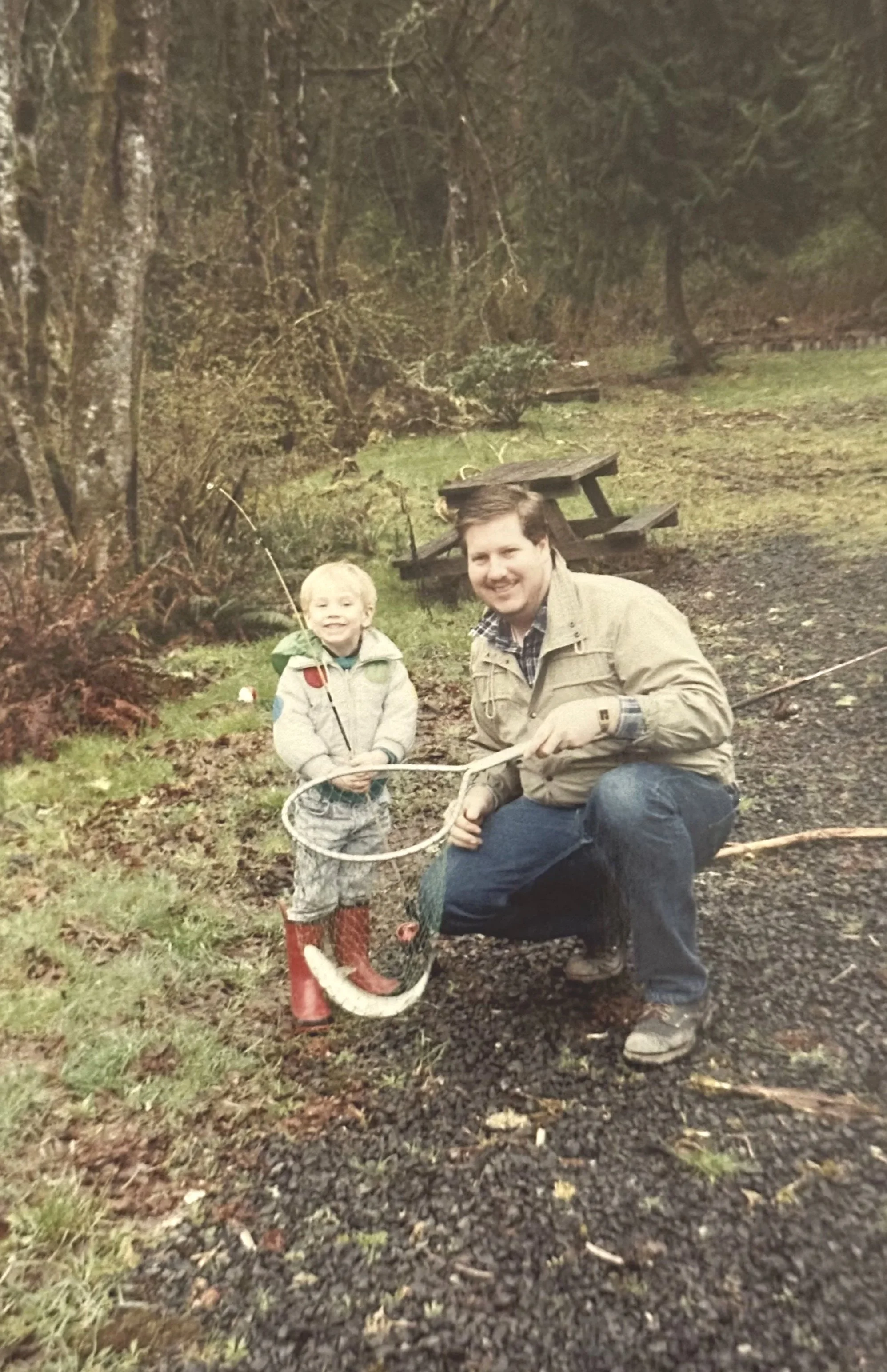 A man and a young boy outdoors, fishing with a net, near a wooded area and a picnic table.