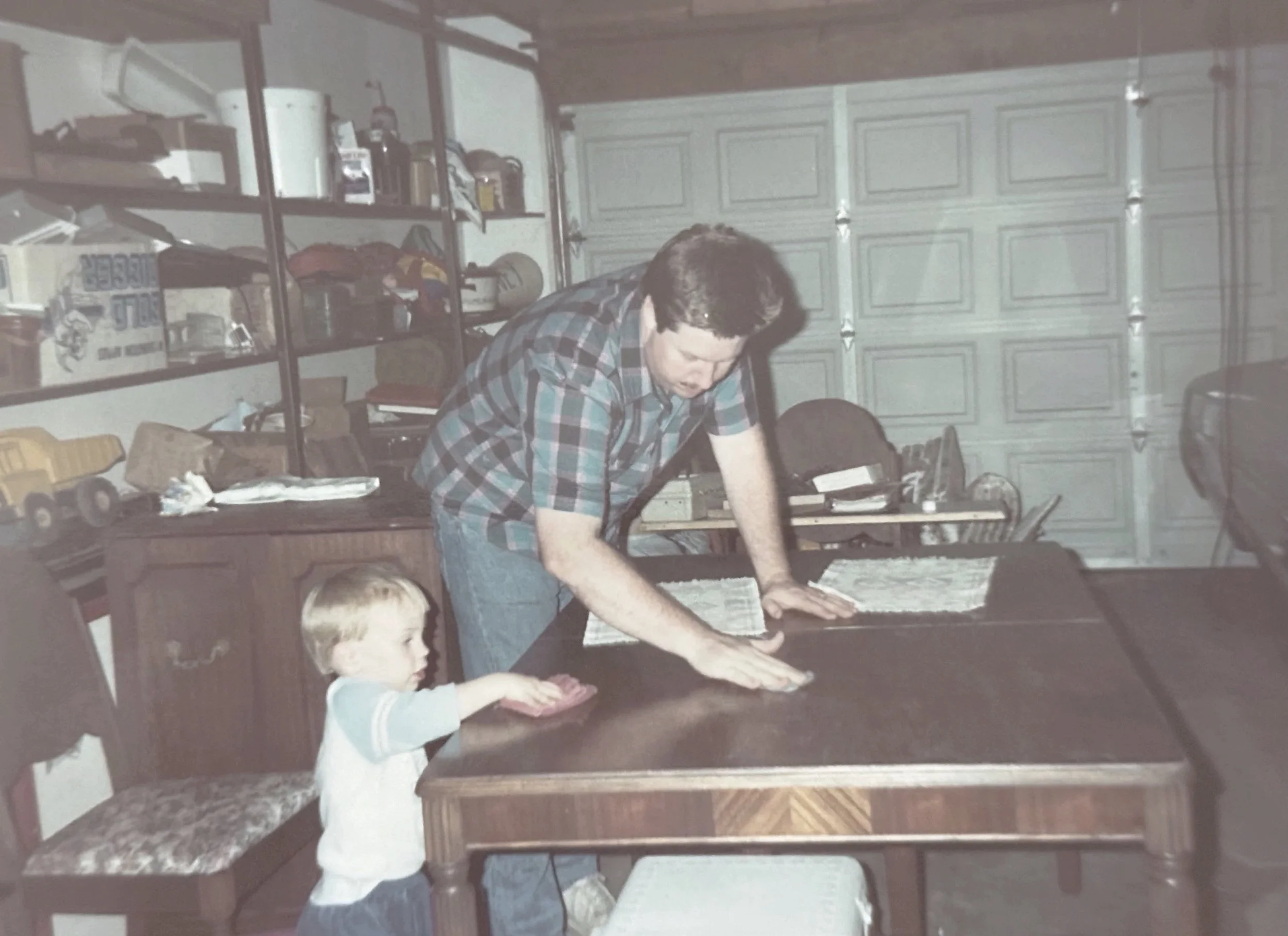 A man in his 30's and a young child cleaning or wiping a dark-colored table in a garage with shelves and a closed garage door in the background.