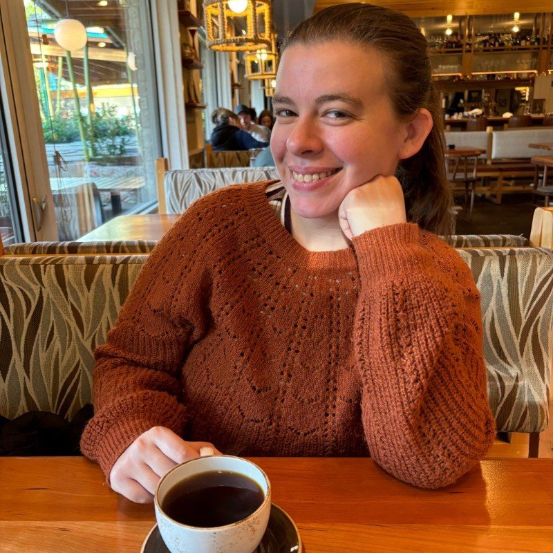 A young woman with brown hair in a ponytail smiling at the camera, seated at a wooden table in a cozy cafe with a cup of coffee in front of her, surrounded by warm decor and other patrons.