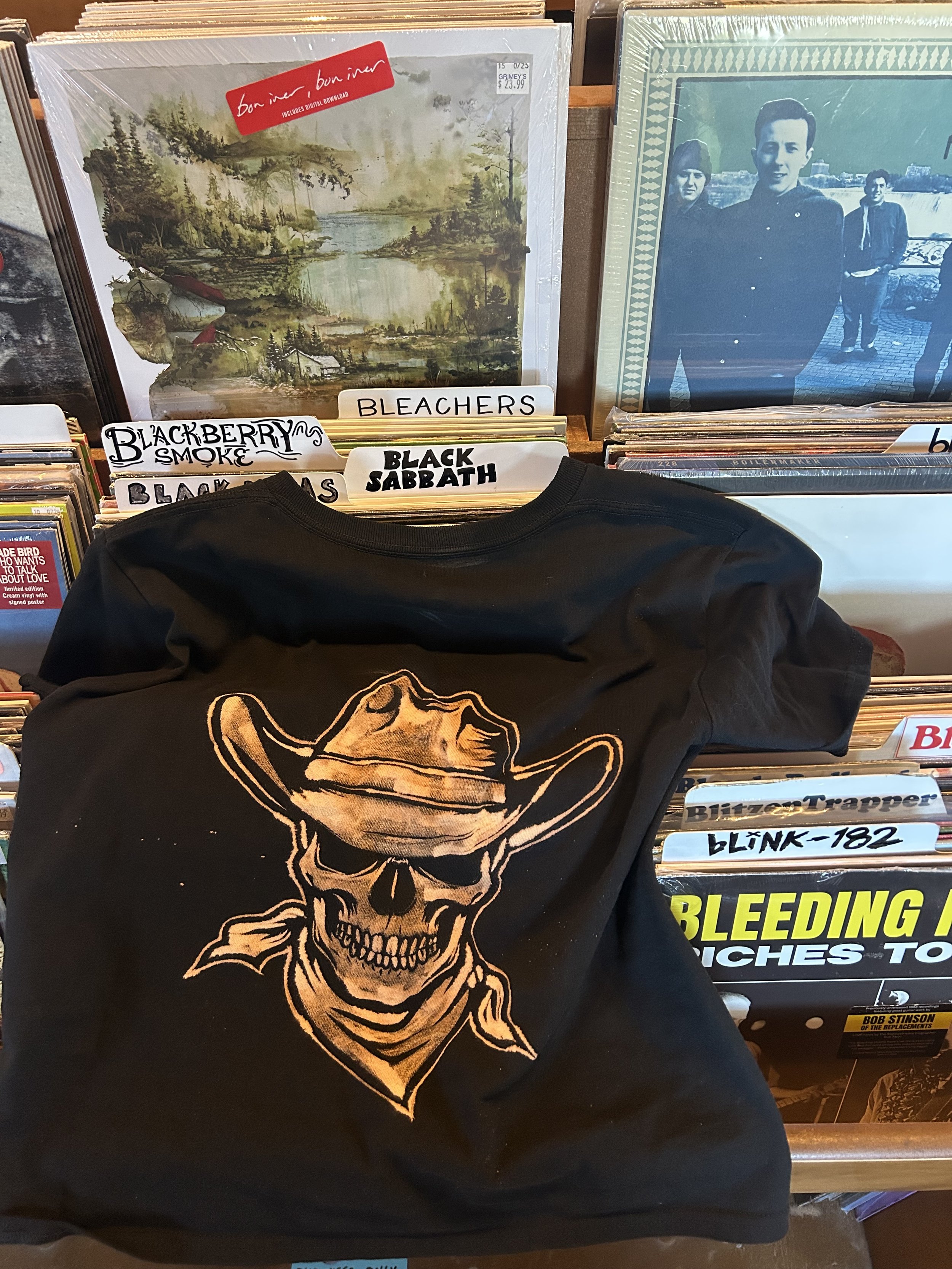Black T-shirt with a graphic of a skull wearing a cowboy hat and bandana, placed on a shelf with vinyl records behind it.