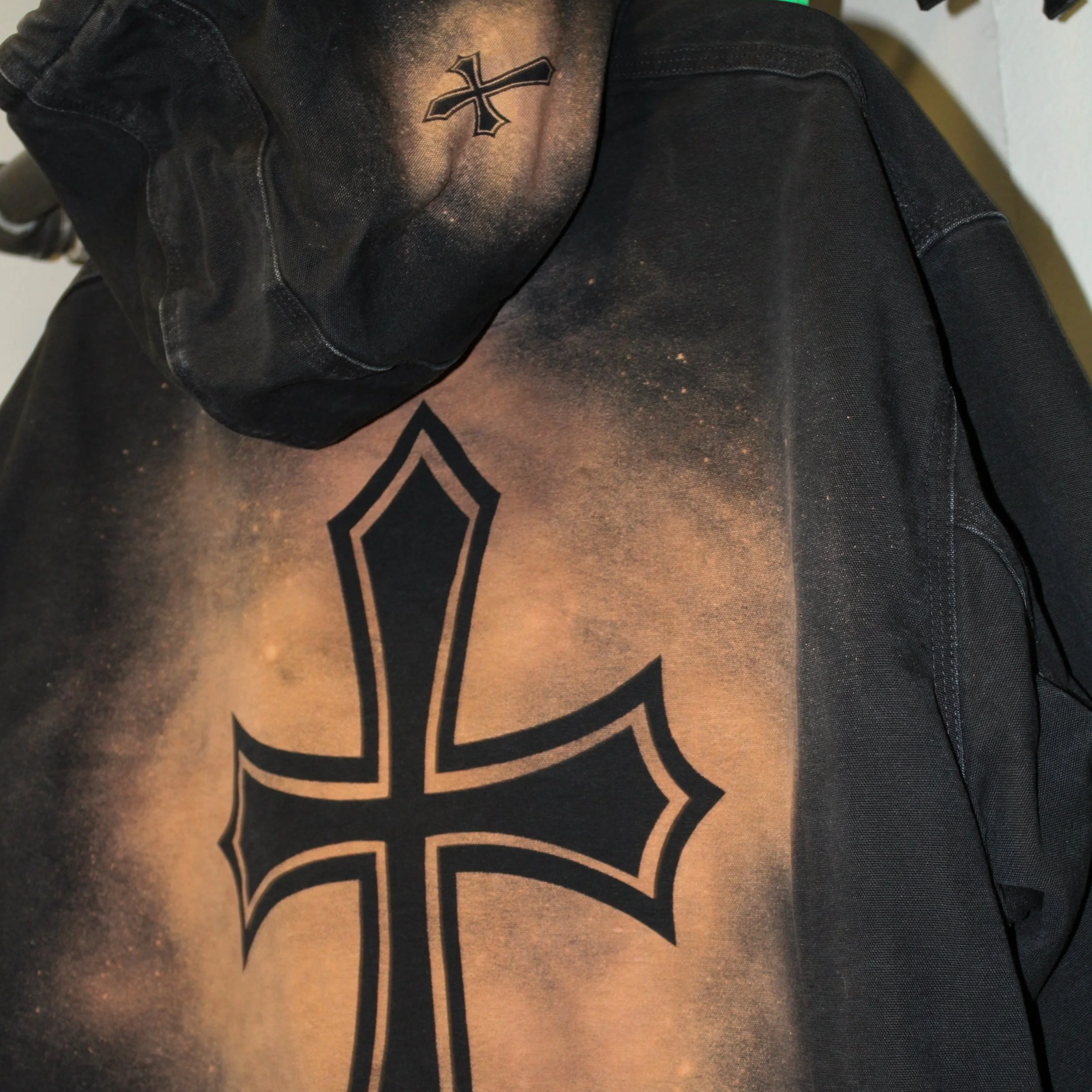 Close-up of a black hoodie with a large cross design and a smaller cross on the hood, with a starry sky and galaxy print in the background.