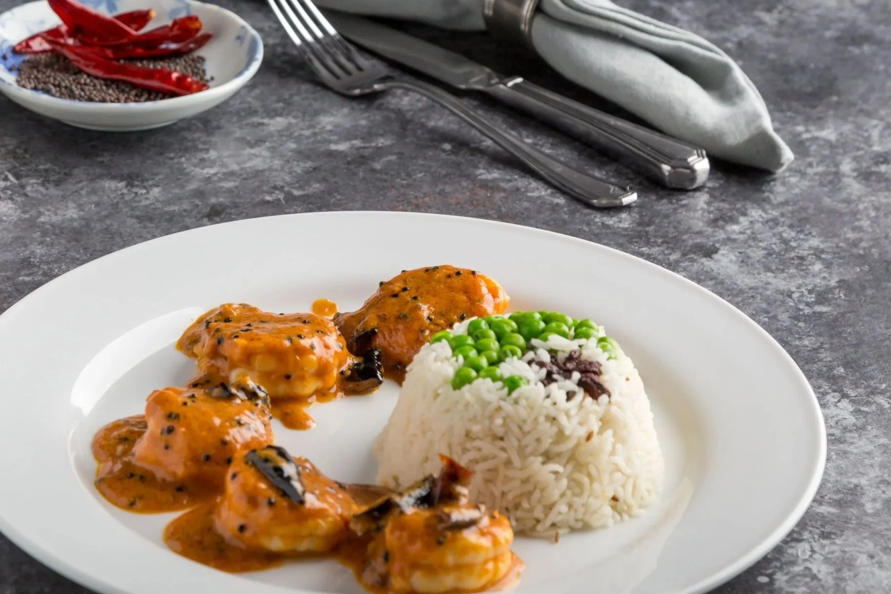 Plate of rice topped with peas and a small amount of sauce, served with three pieces of shrimp covered in a spicy orange sauce, on a white oval plate. In the background, a bowl with dried red chili peppers and black sesame seeds, along with a fork, knife, and gray napkin on a dark gray countertop.