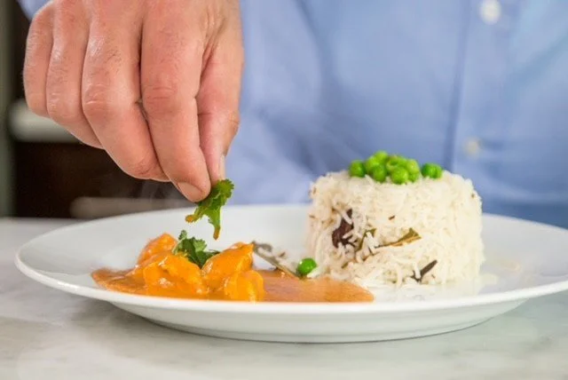 A person garnishing a plate of food with cilantro, with a serving of rice topped with peas and a side of chicken in sauce.