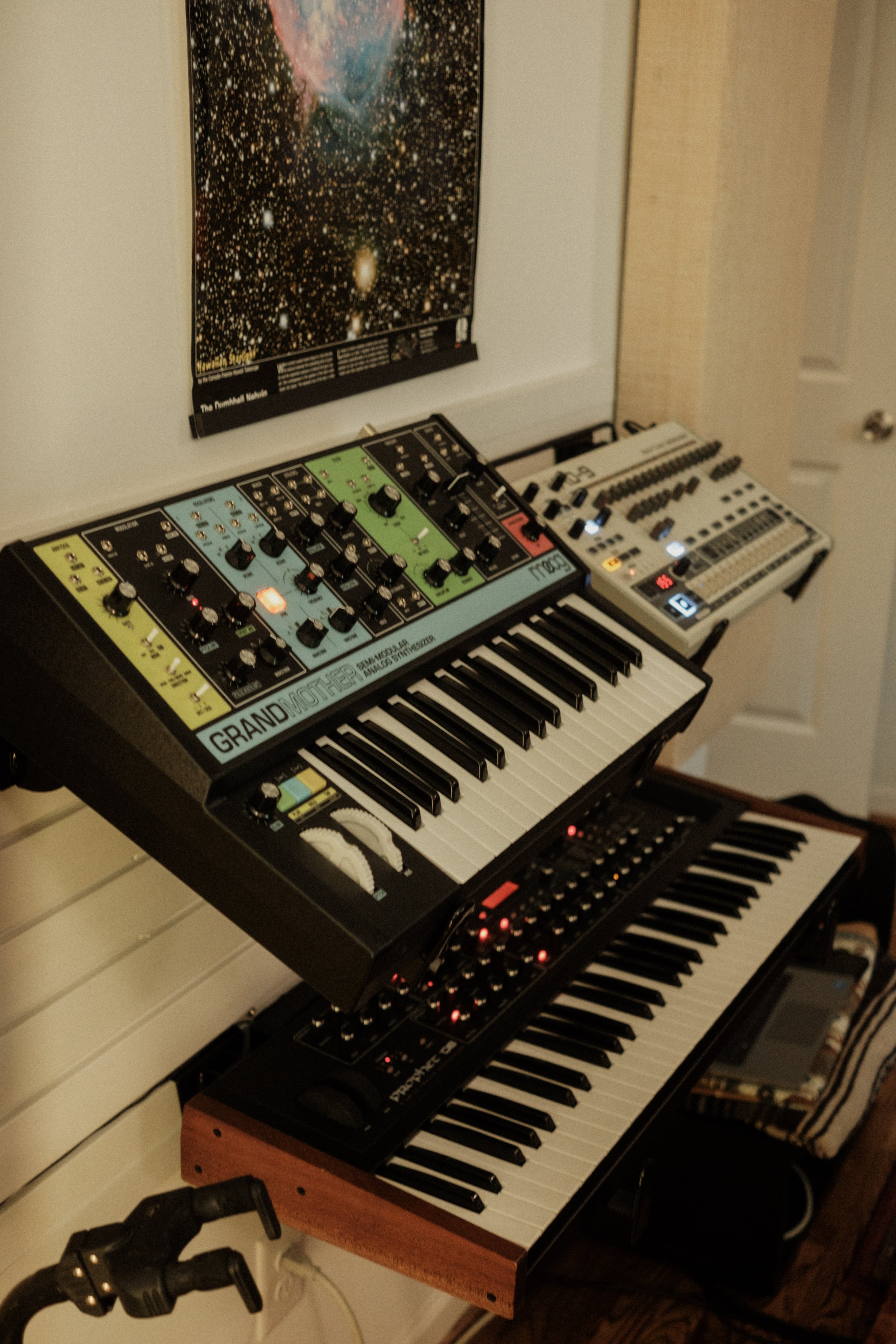 Photo of a music studio setup with two synthesizers, a keyboard, and an electronic drum module, located near a wall with a galaxy-themed poster.