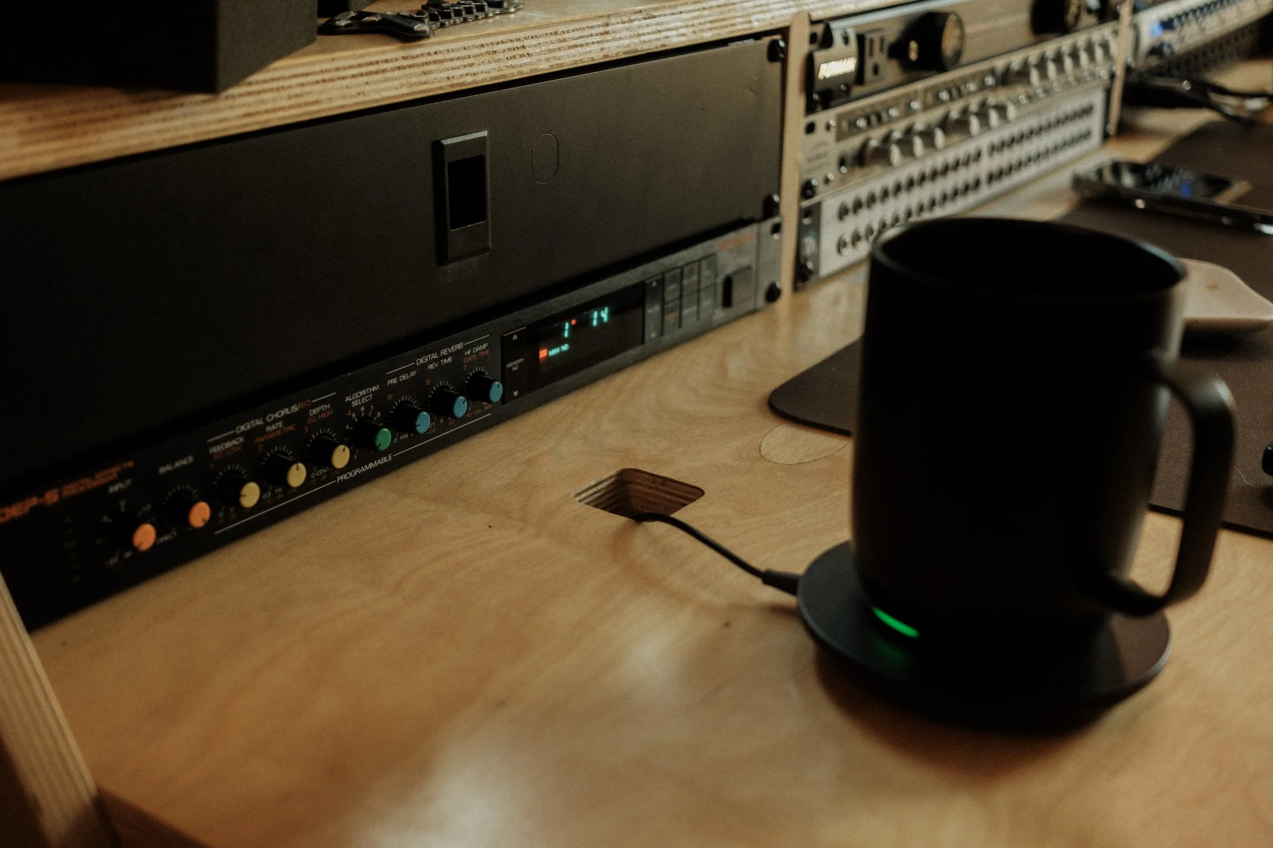 An electronic music workstation setup on a wooden desk with audio equipment, computer accessories, and a black mug on a coaster.