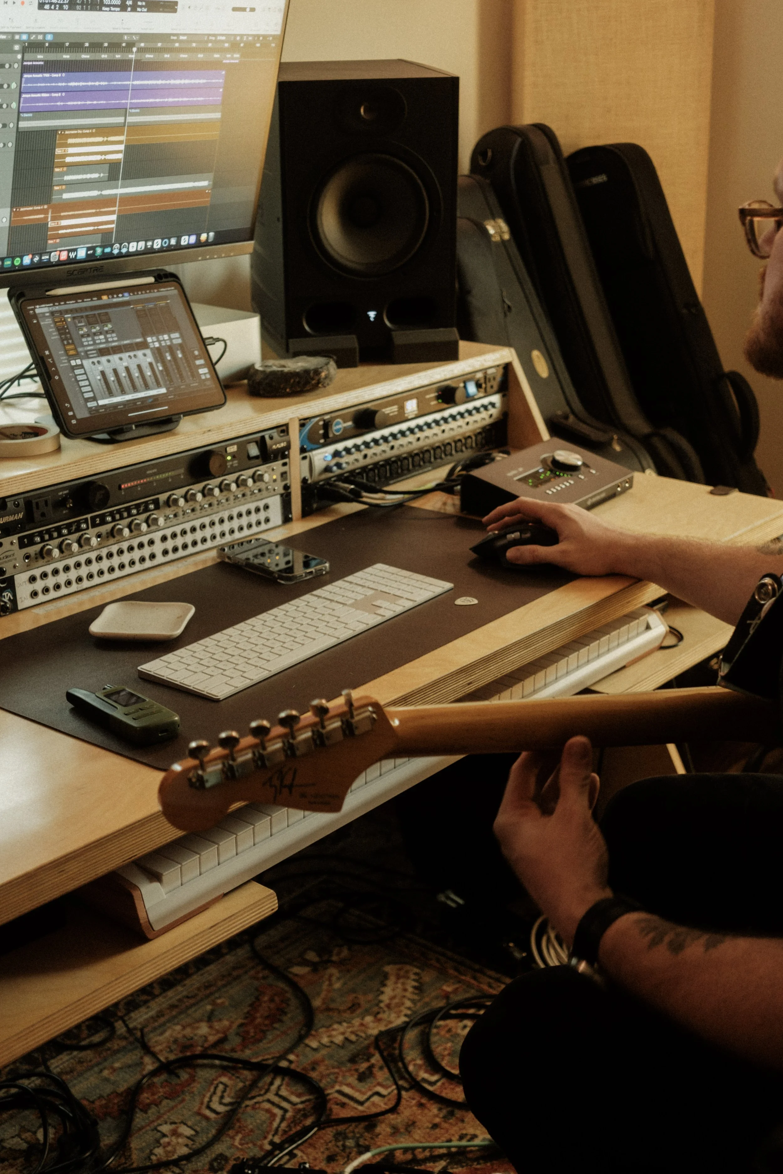 A person working in a music studio with a keyboard, computer, keyboard controller, tablet, studio monitor speakers, a guitar, and various audio equipment, including a mixer and audio interfaces.