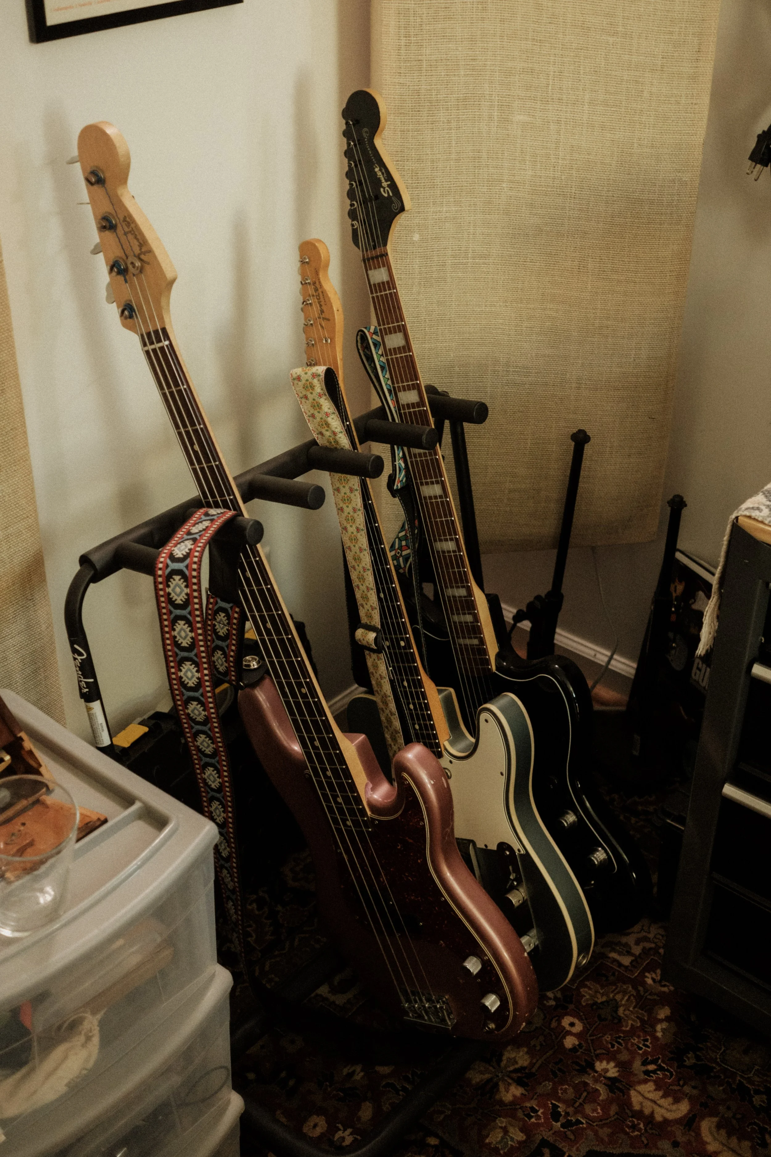 Four guitars resting on guitar stands in a room, with a small drawer unit and a patterned rug visible.