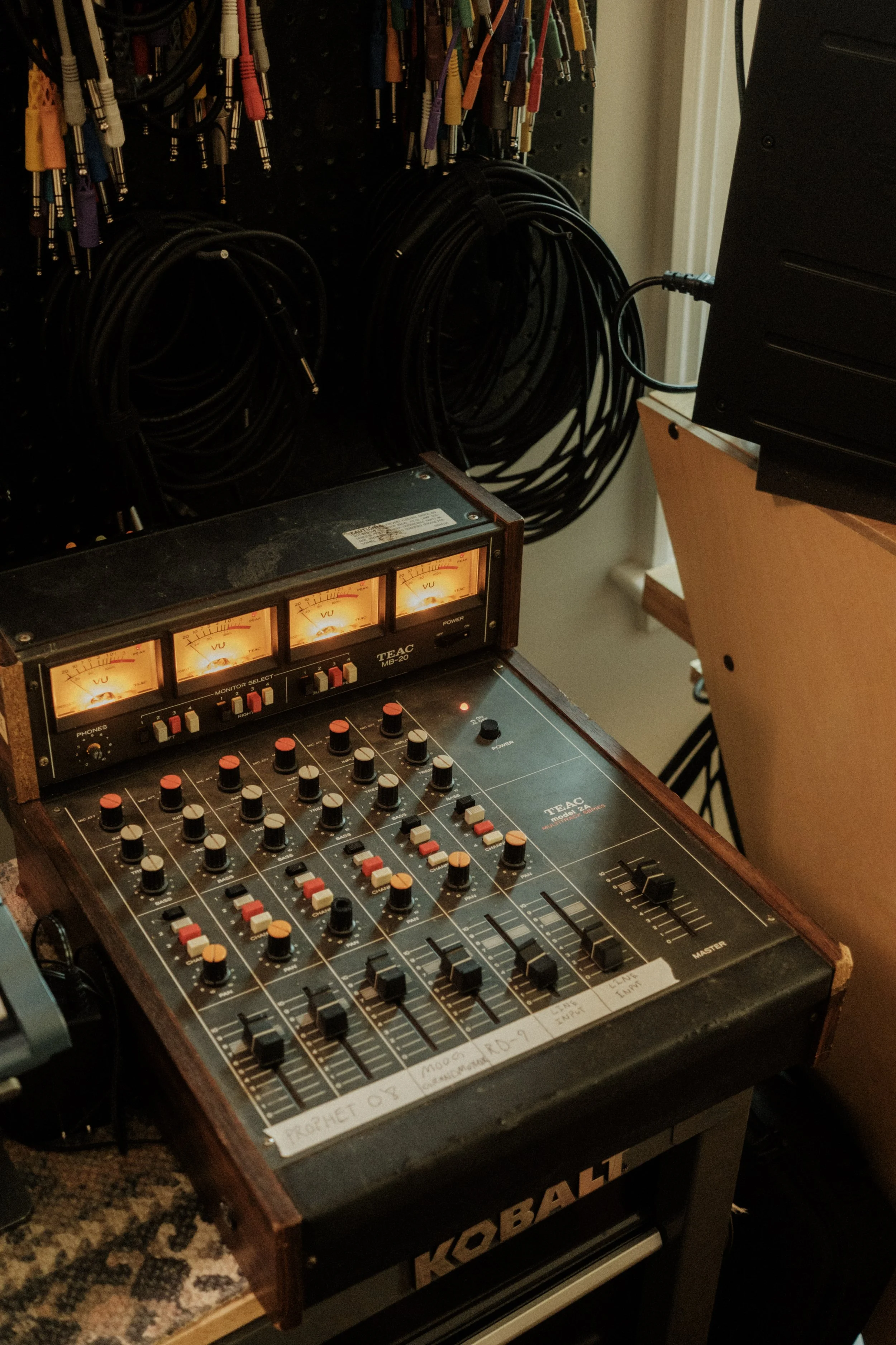 A vintage audio mixing console with several illuminated meters, numerous knobs, and sliders, labeled 'KOBALT.' Behind the console, there are coiled cables hanging on a pegboard and some equipment on a side table.