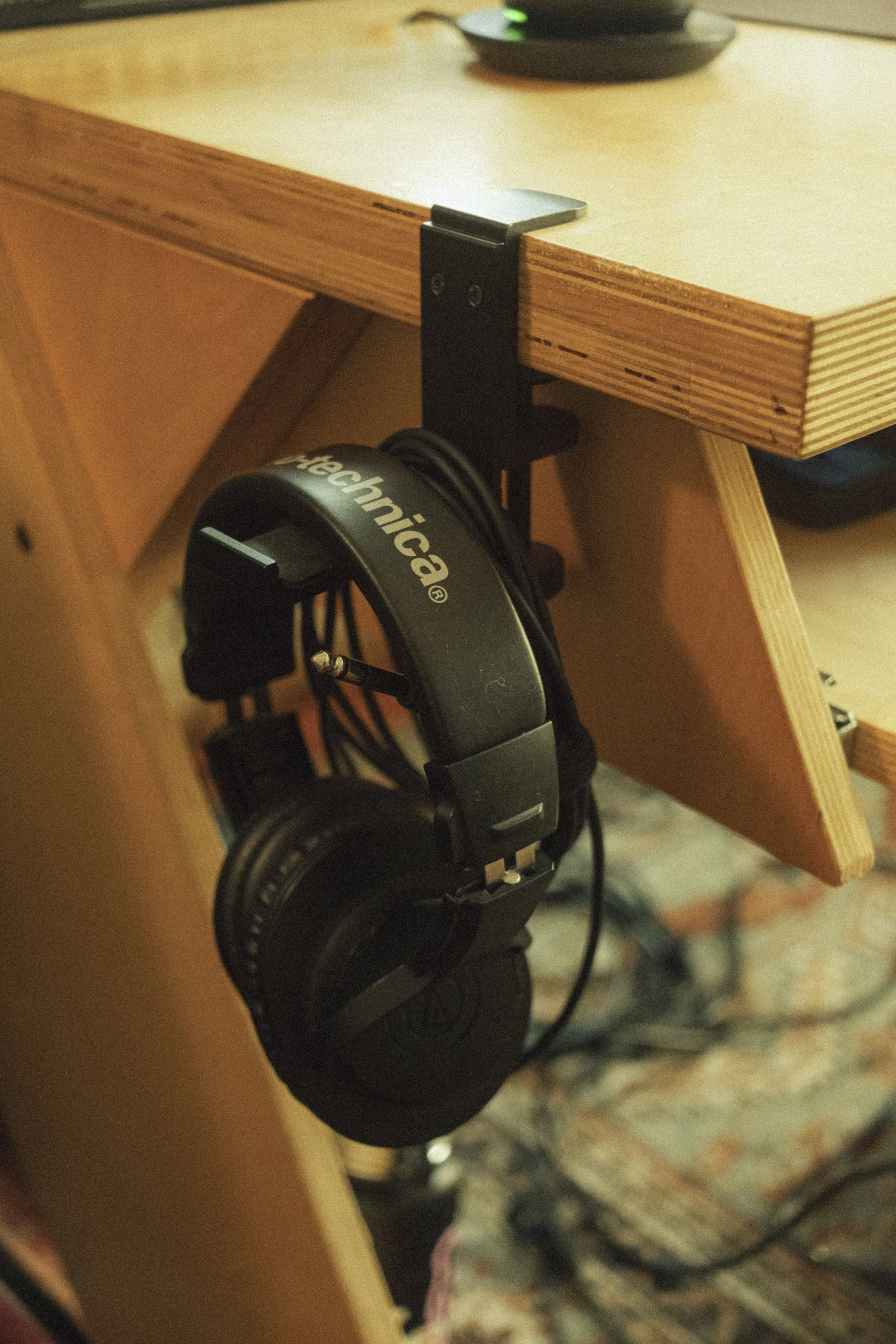 A pair of black headphones hanging from a hook under a wooden desk.