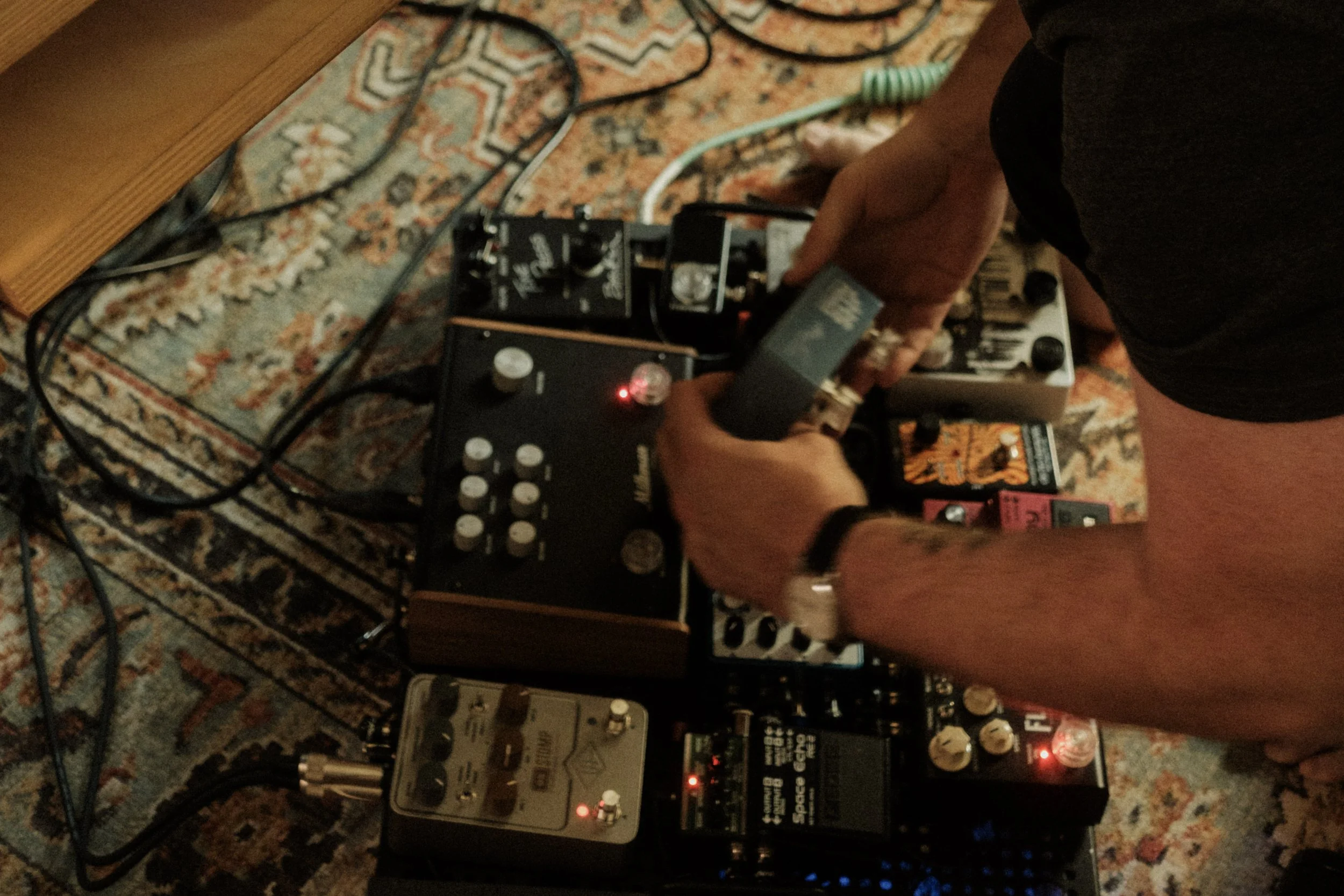 A person operating a guitar effects pedalboard setup on a carpeted floor, with multiple effects pedals, cables, and audio equipment.
