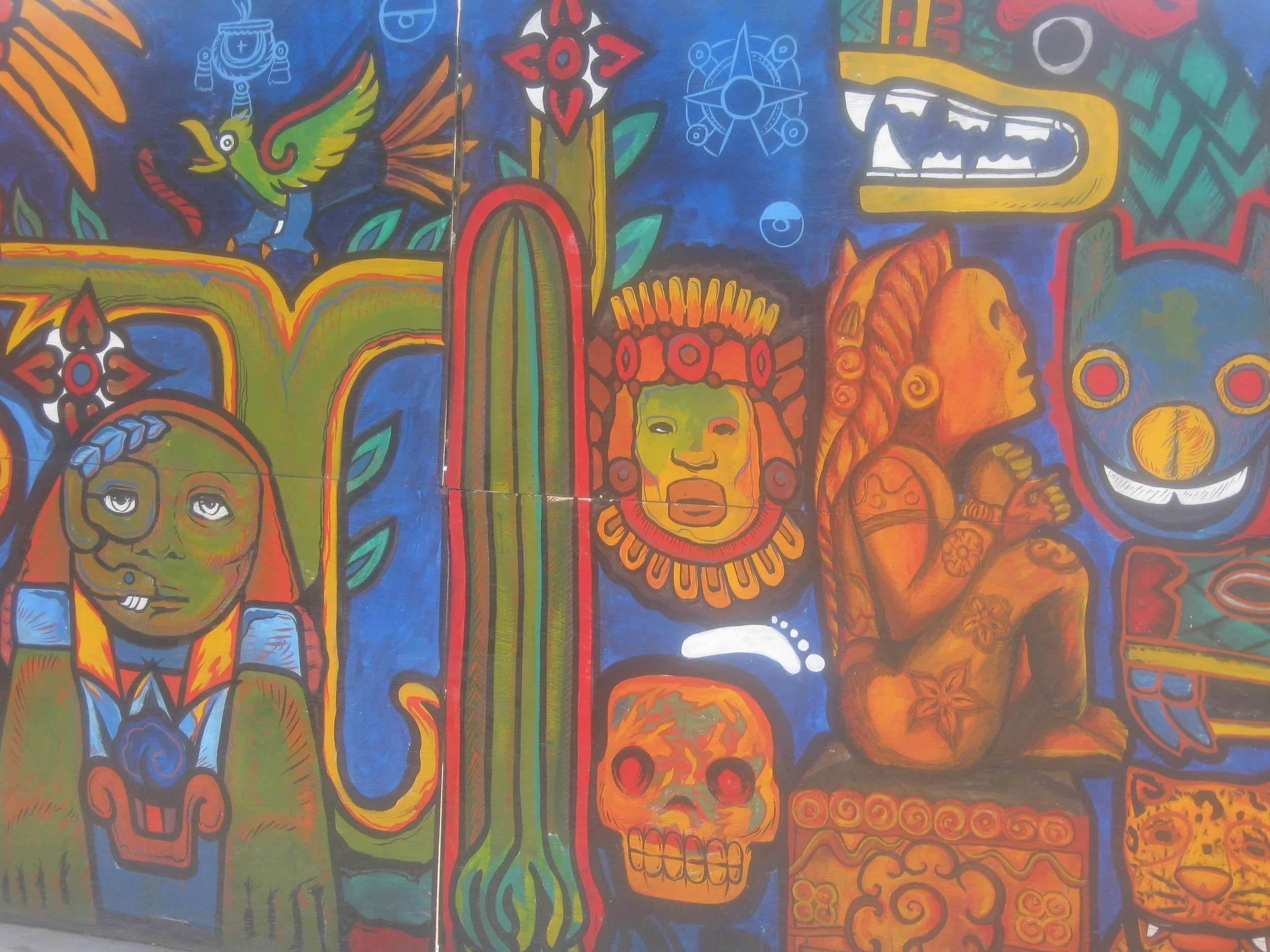Colorful mural with various indigenous and cultural images, including stylized faces, a skull, a brightly painted snake, a person with features resembling a totem or mask, and a bird. The mural uses vivid reds, blues, greens, and oranges.