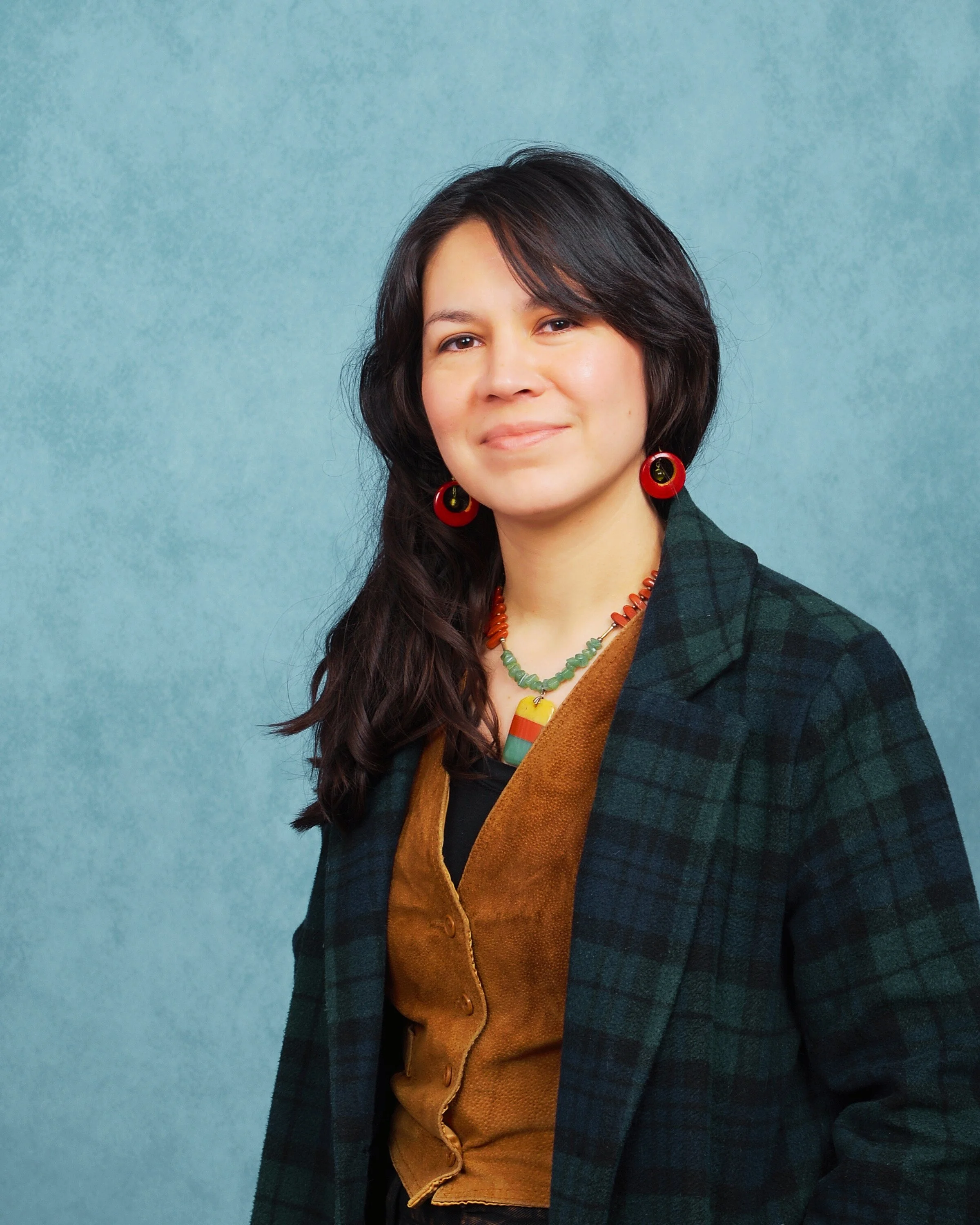 A woman with shoulder-length dark hair wearing red earrings, a colorful beaded necklace, a black top, a green plaid blazer, and a brown vest stands against a textured blue background.