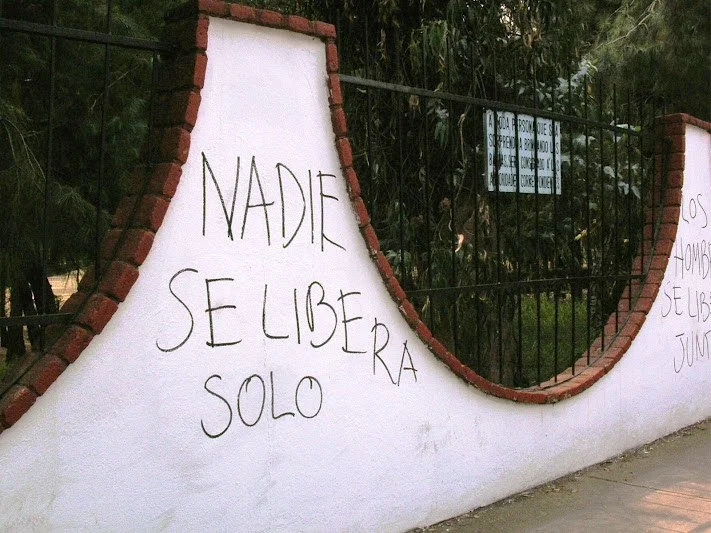 White wall with red brick trim and black metal fence, with graffiti in Spanish that reads 'Nadie se libera solo'.