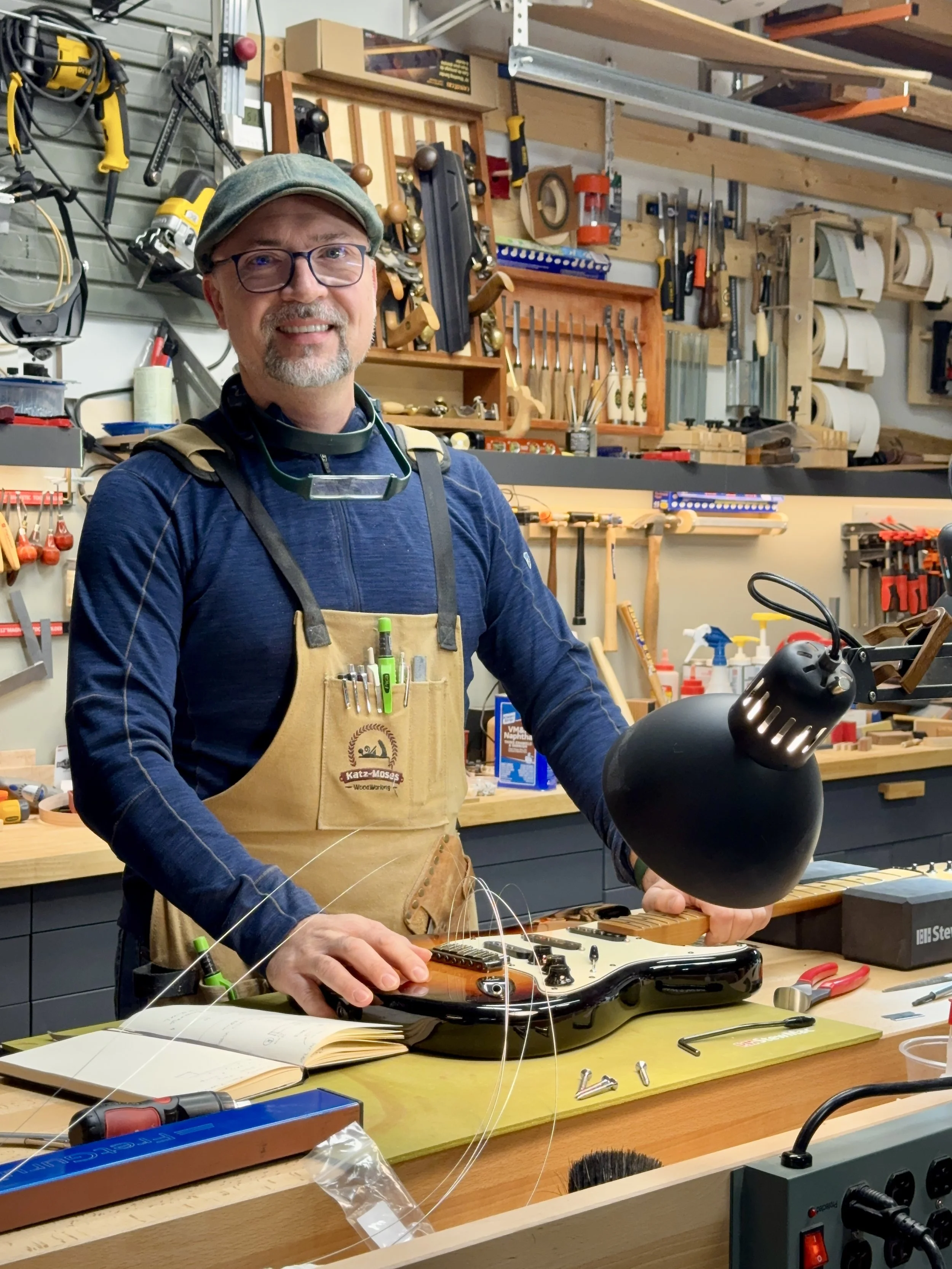 A man in a woodworking workshop holds a black lamp and an electric guitar with wires on a workbench.