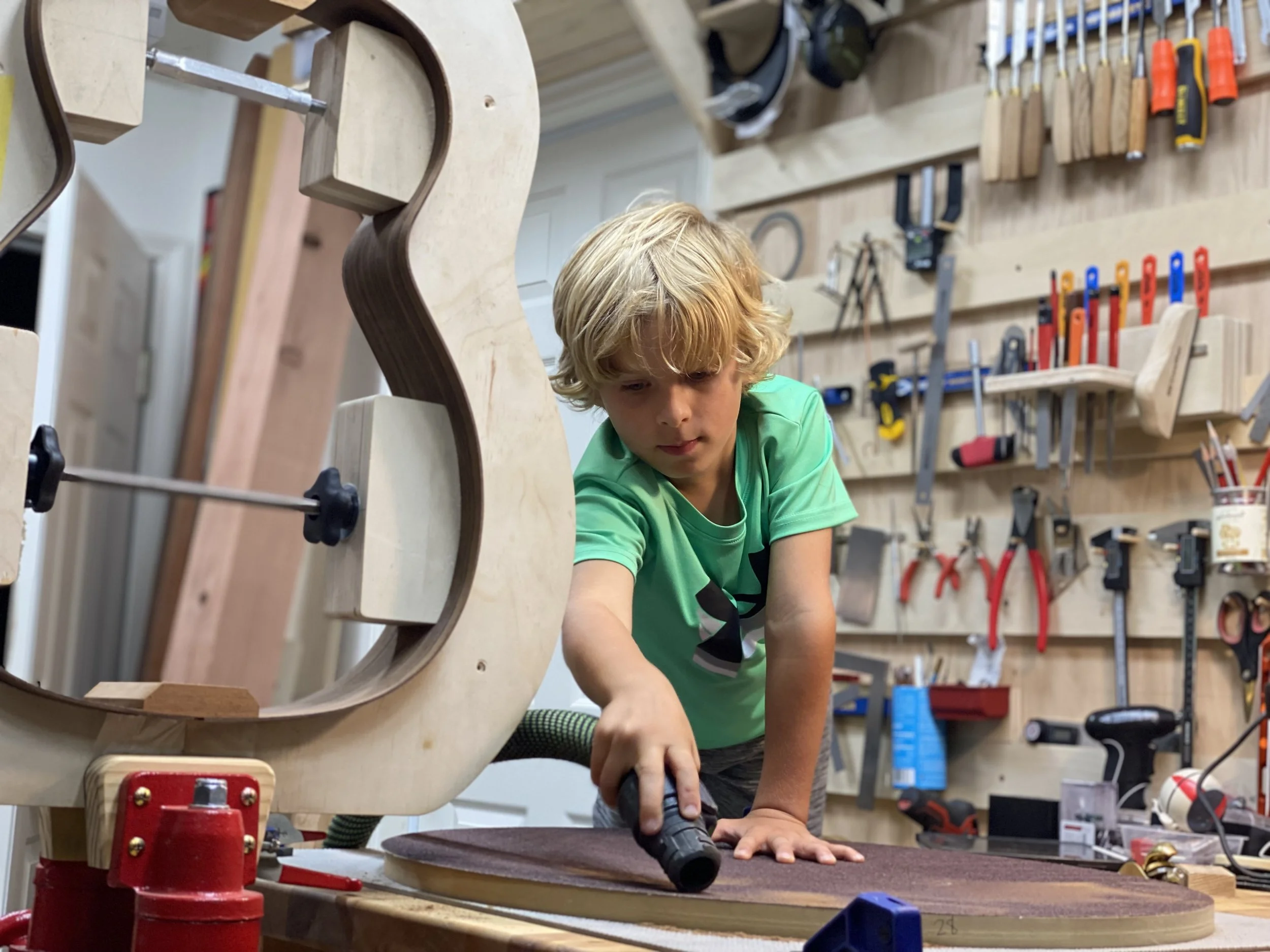 A young boy with blonde hair, wearing a green T-shirt, is using a handheld power tool on a piece of wood in a woodworking workshop. The background shows various hand tools and equipment hanging on the wall.