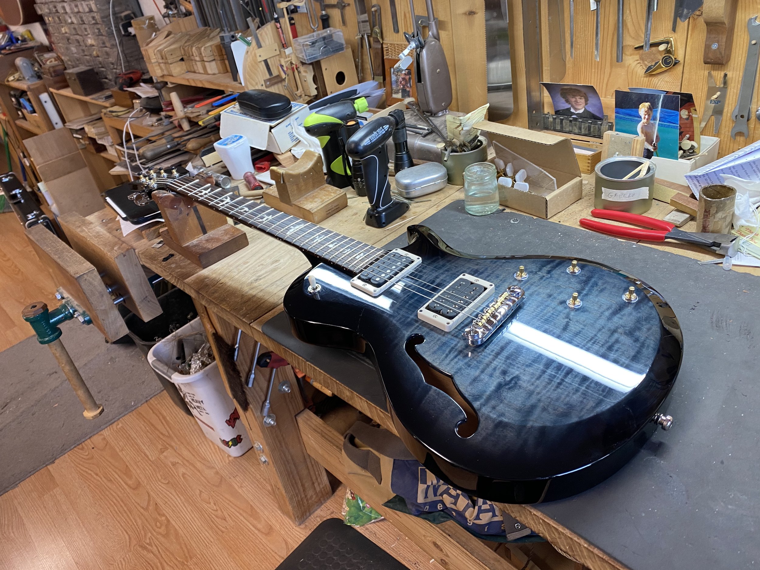 A blue electric guitar on a workbench surrounded by woodworking tools and photographs in a workshop.
