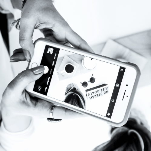 A person is taking a photo of a flat lay arrangement with a smartphone. The arrangement includes a coffee cup, sunglasses, a camera, and a sign that reads 'NO DREAMS JUST GOALS.'