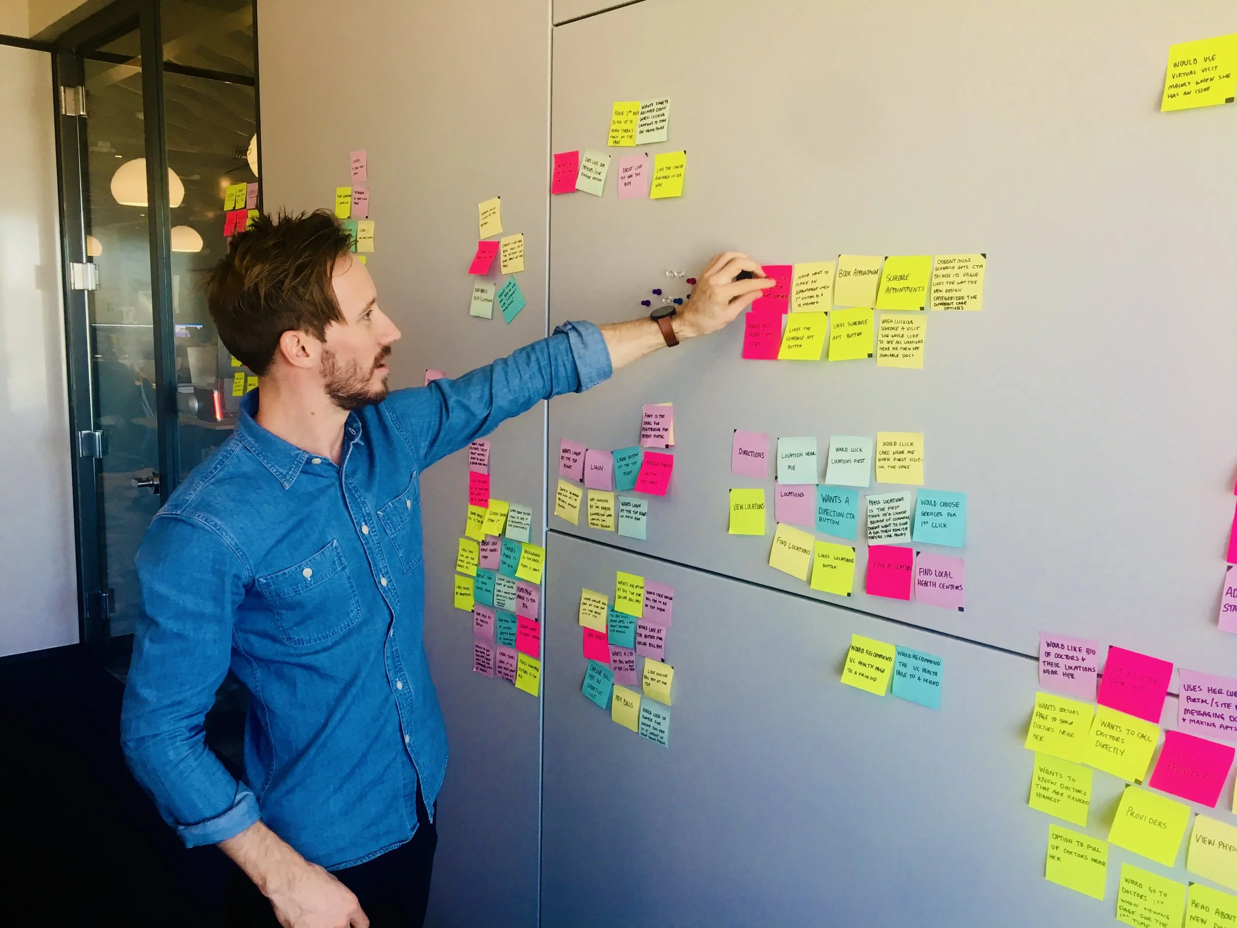 A man with brown hair and beard, wearing a blue denim shirt, standing front of a gray wall with colorful sticky notes arranged on it, likely brainstorming or organizing ideas.
