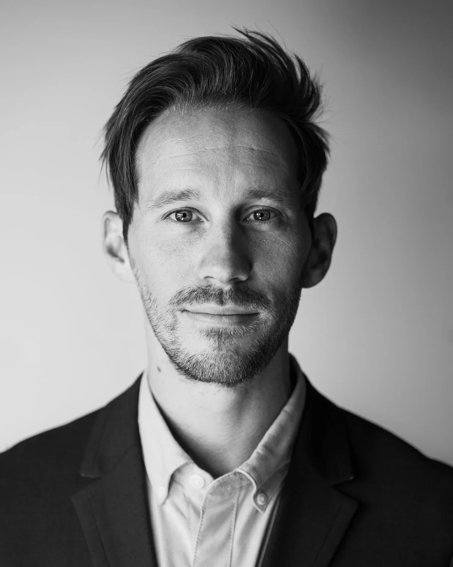 Black and white portrait of a young man with short hair, slight beard, wearing a suit jacket and white collared shirt, looking directly at the camera with a neutral expression.