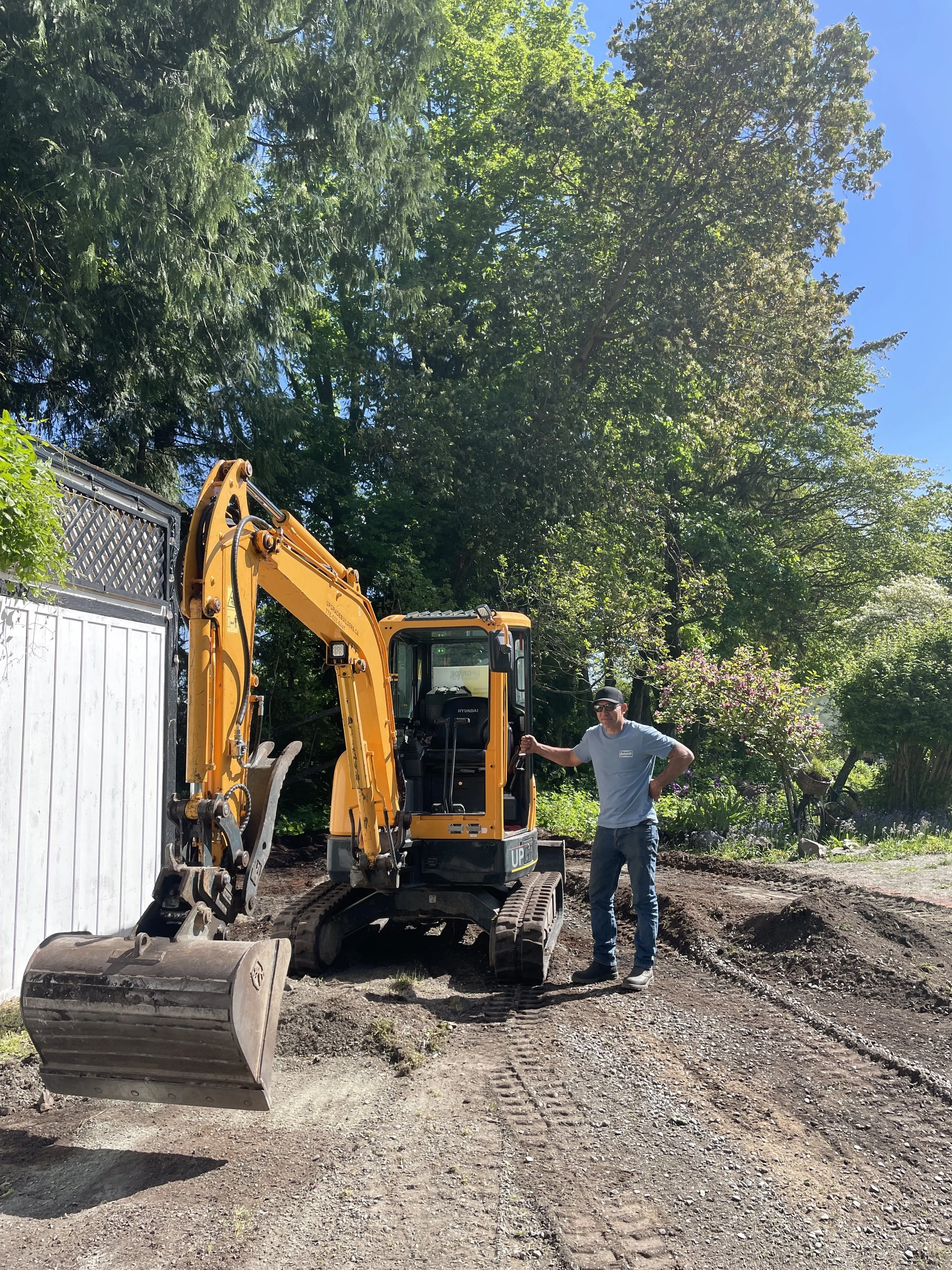 Owner of Upgrade Builders Inc stands next to a small yellow excavator on a dirt path, surrounded by green trees and a blue sky on Vancouver Island.