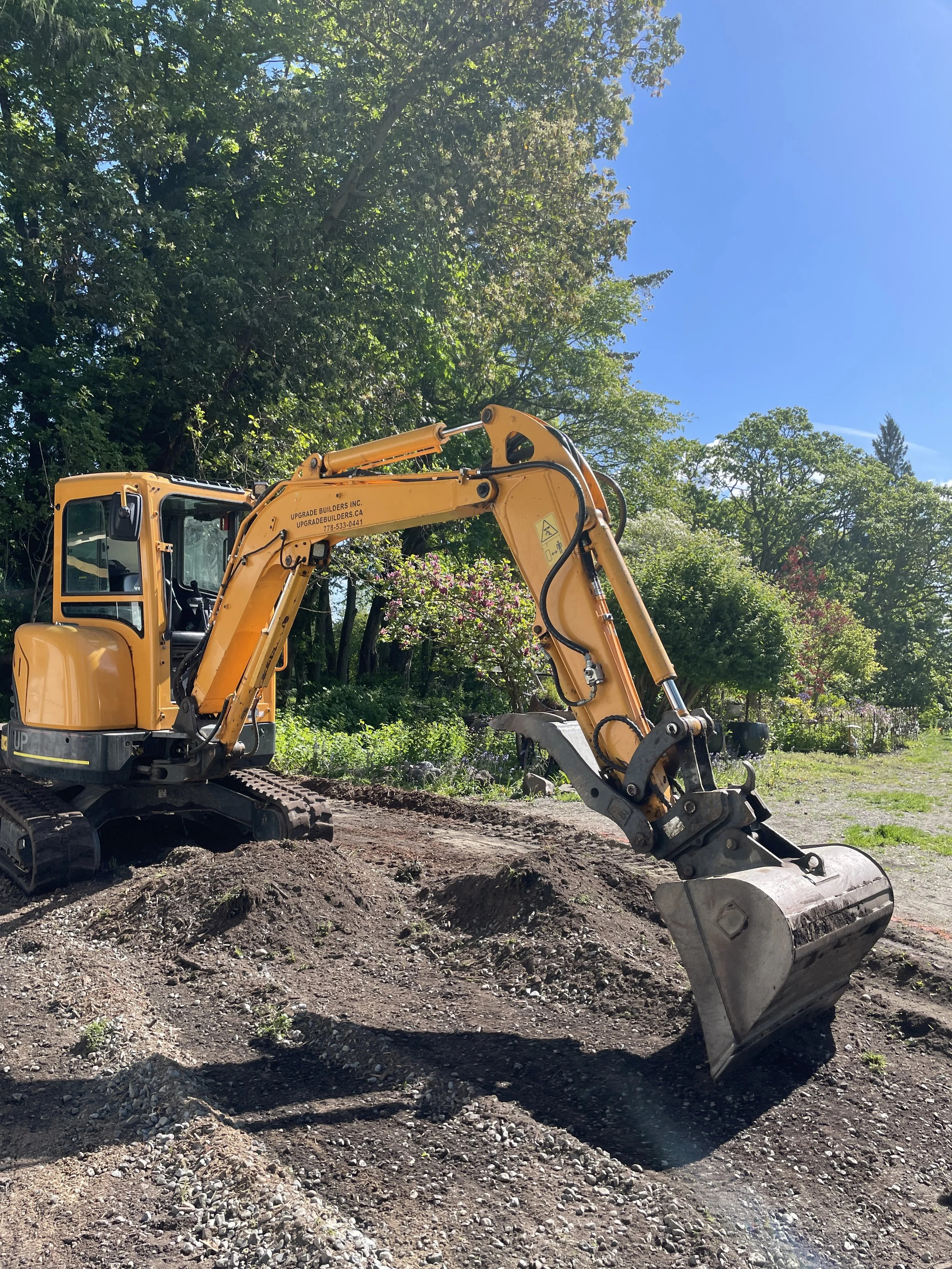 A yellow mini excavator digging in a dirt to create a new driveway, with trees and blue sky in the background.