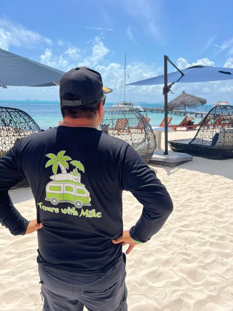 Man wearing black cap and black shirt with green palm tree and camper van graphic, standing on a sandy beach facing water with lounge chairs and umbrellas, and boats docked at a pier in the background.