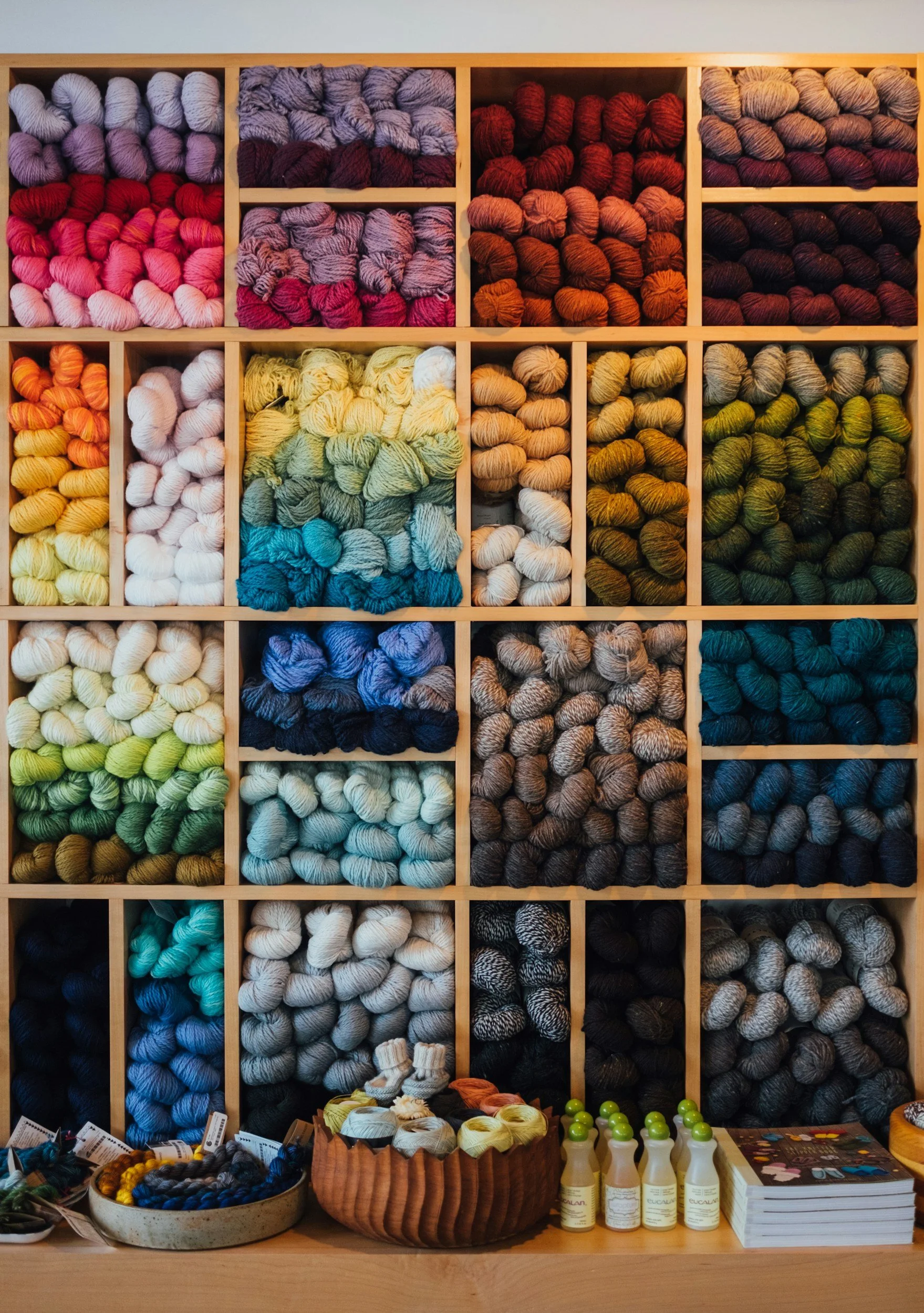 Colorful yarns arranged in a display shelf, with various skeins in shades of pink, purple, red, orange, yellow, green, blue, and gray, and small bottles and books on a wooden table in front.