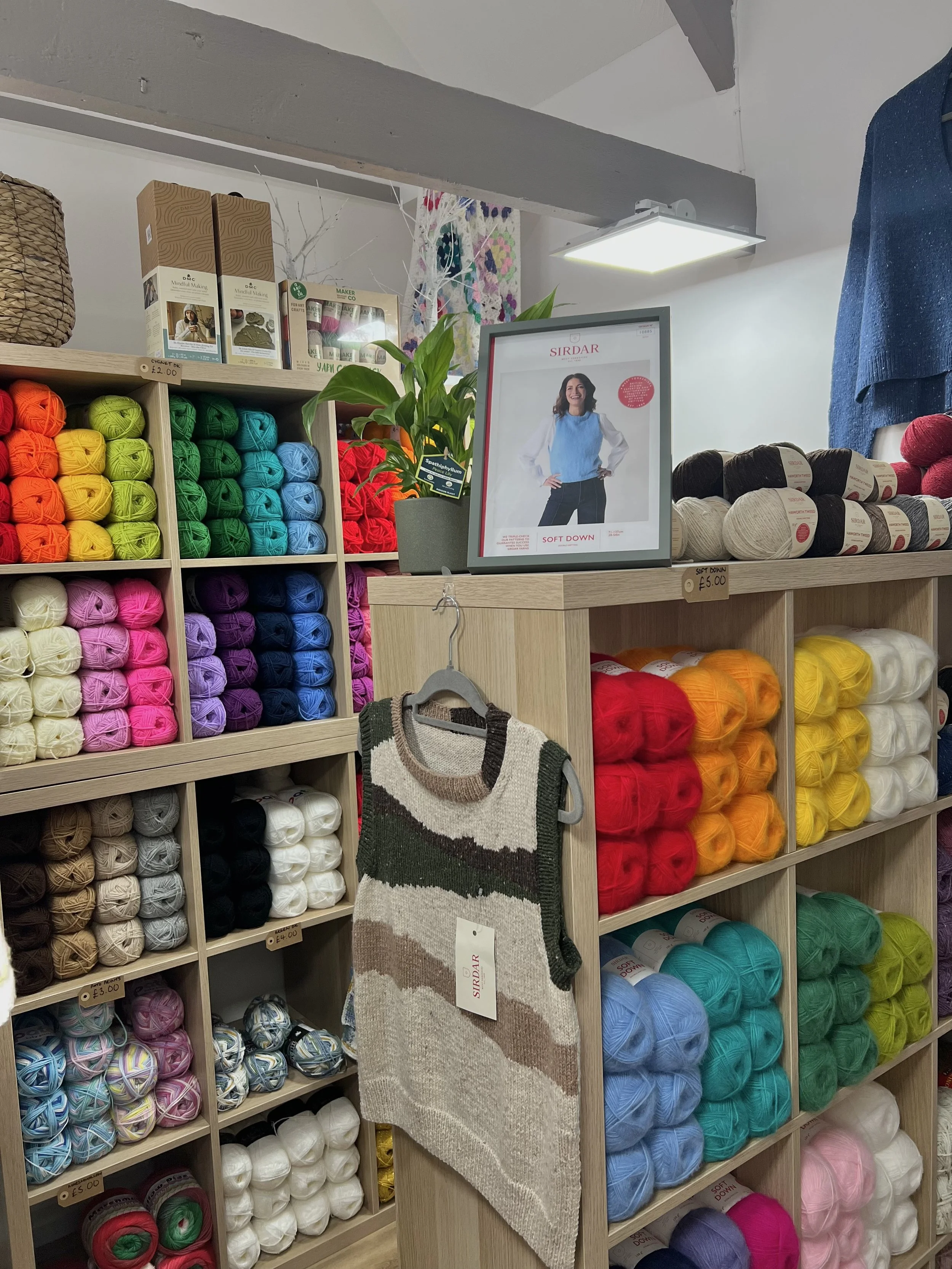 Display of colorful yarns in a craft store, with a framed advertisement for Sirdar yarn and a sleeveless sweater on a hanger.