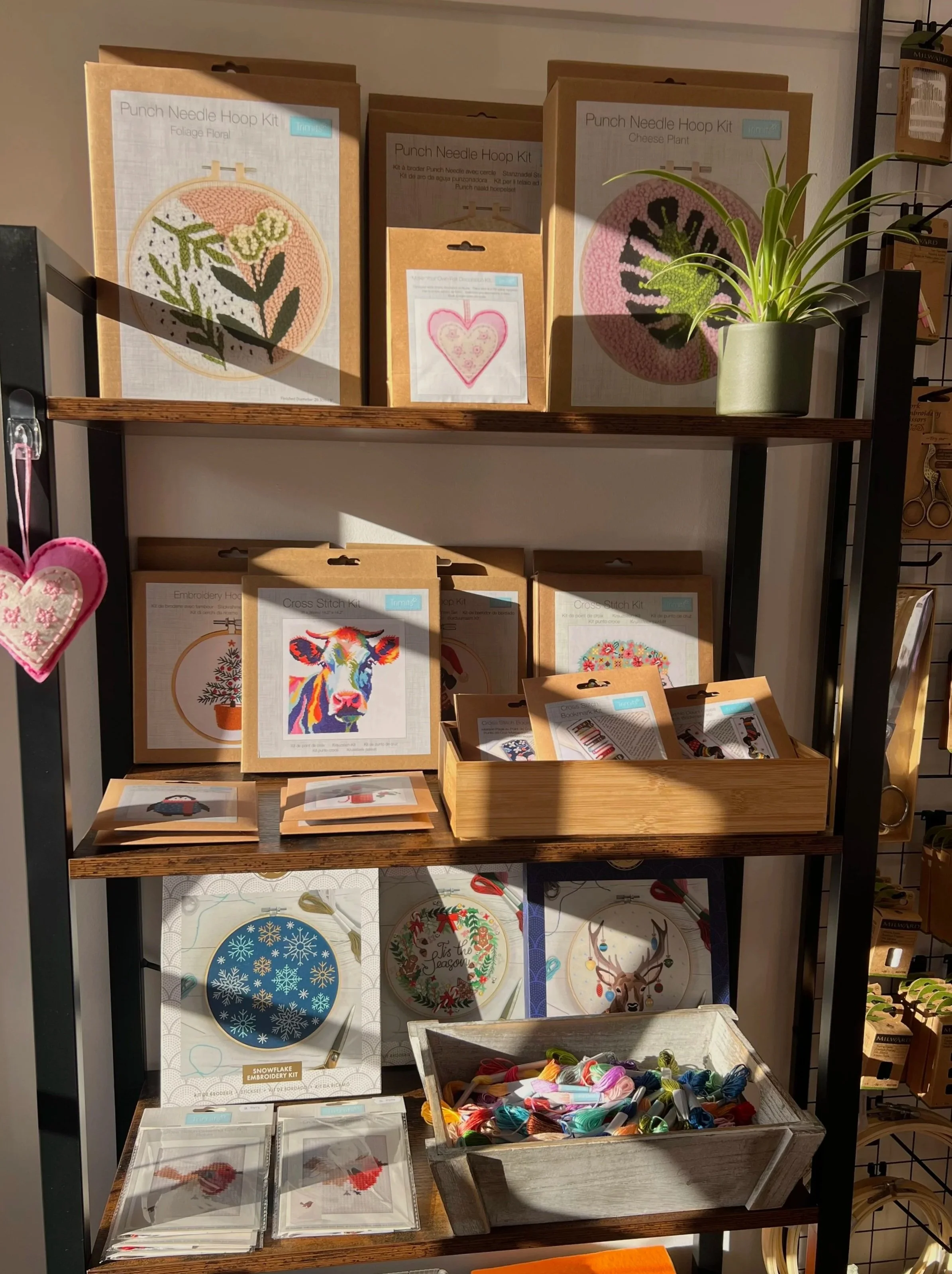 Display shelf with various embroidery kits, cross-stitch sets, and accessories, including punch needle hoop kits, Christmas-themed embroidery, and colorful thread spools.