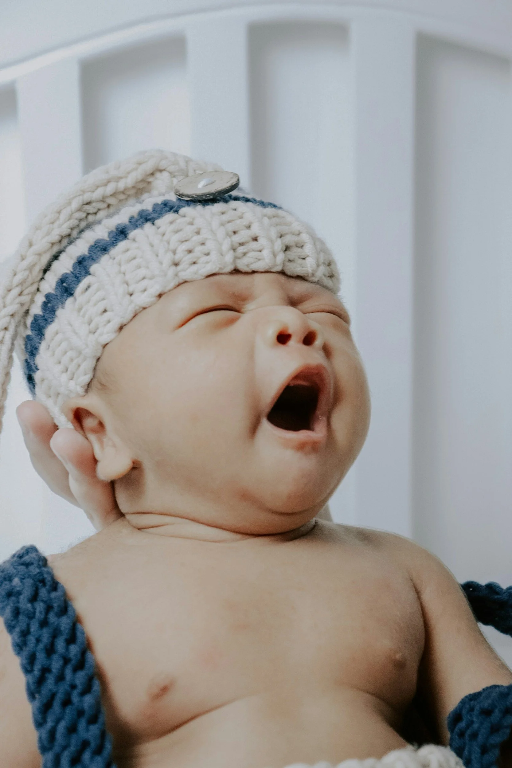Close-up of a yawning baby with a knitted hat being held in someone's hand.