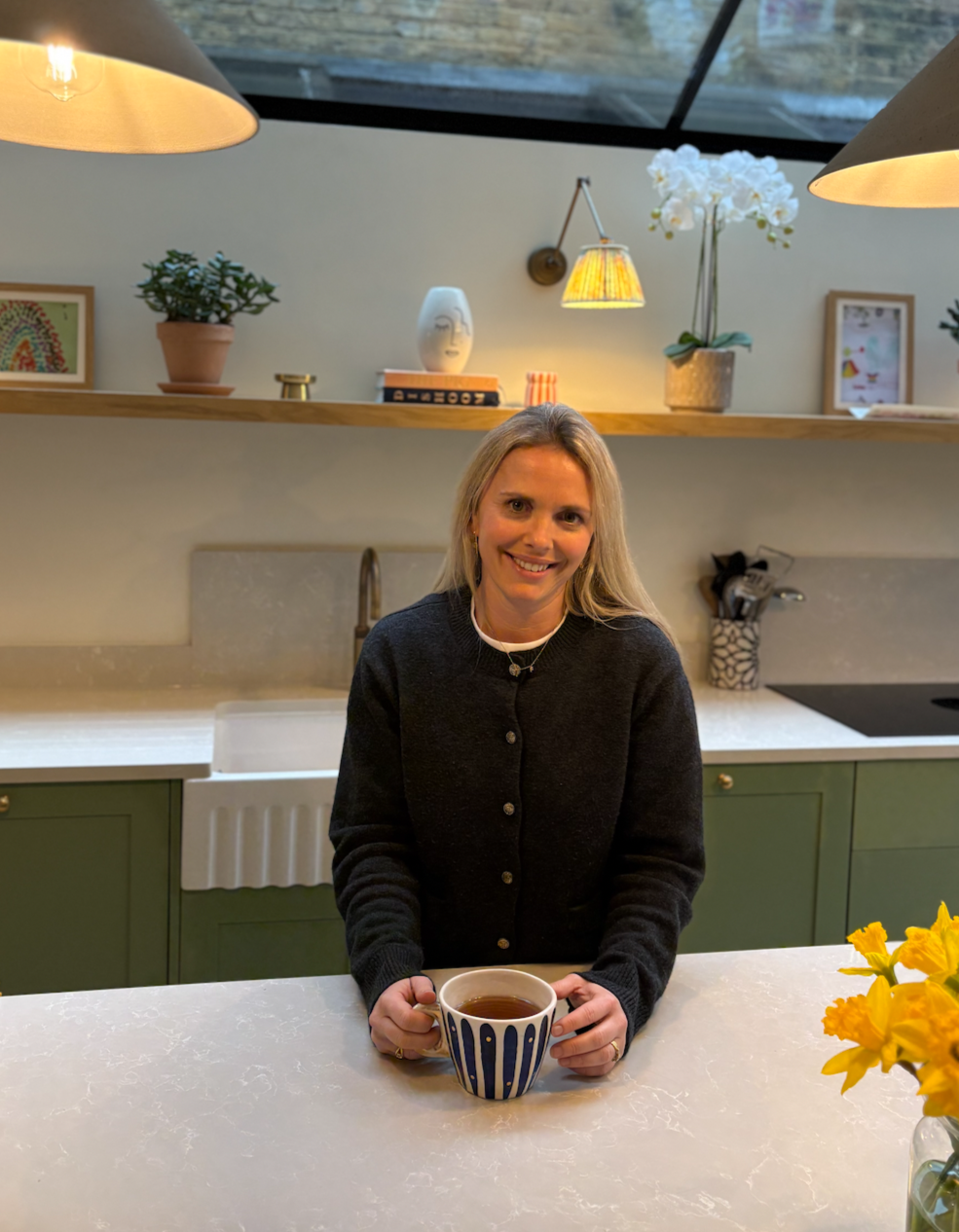 A woman with blonde hair sitting at a kitchen counter, holding a cup of tea or coffee, smiling at the camera. The background features a modern kitchen with green cabinets, a white sink, decorative items, a potted plant, and a vase with white orchids, illuminated by warm lighting.