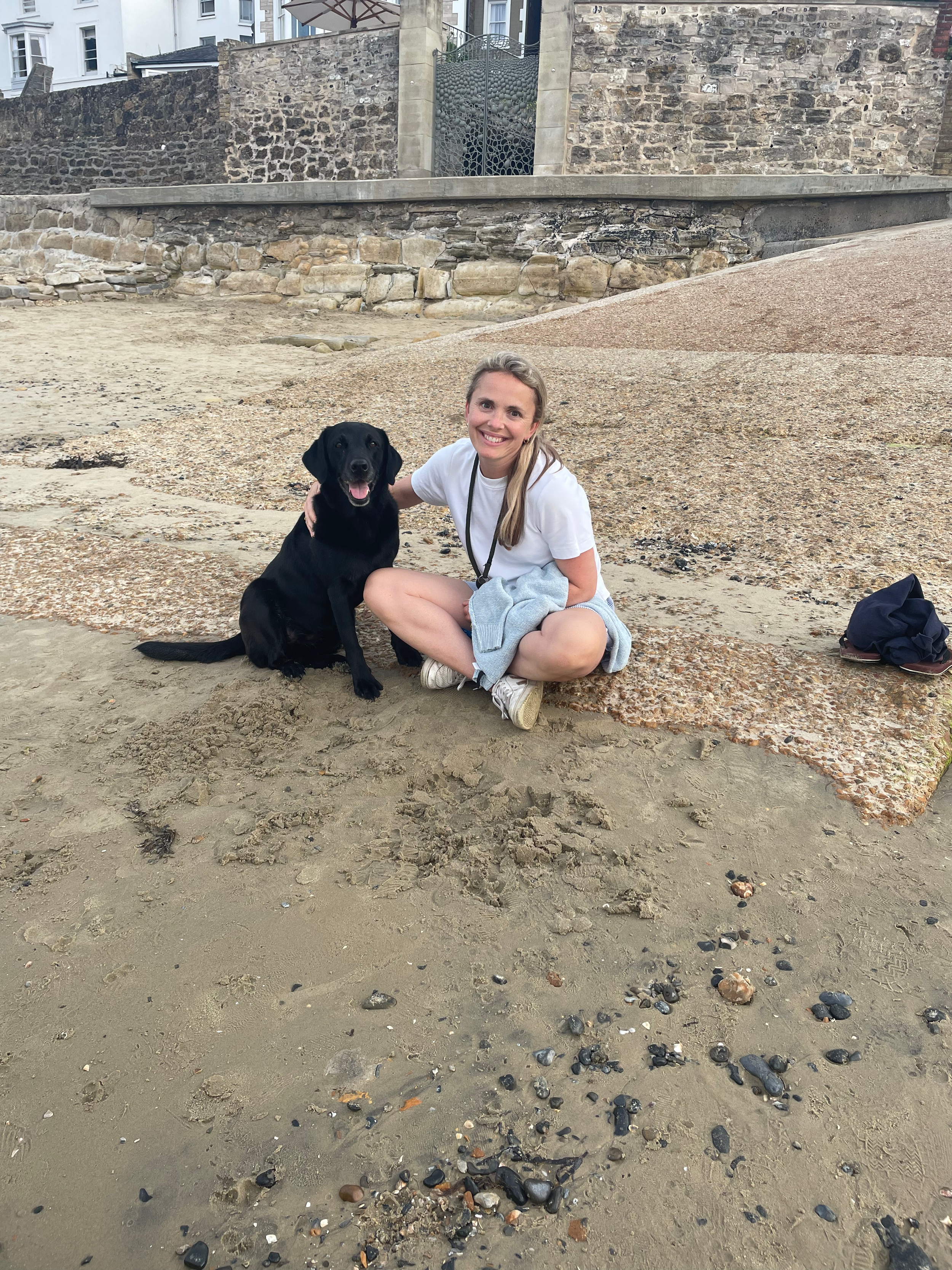 A woman sitting on a sandy beach next to a black Labrador Retriever dog, both smiling. Behind them is a rocky wall and brick buildings.