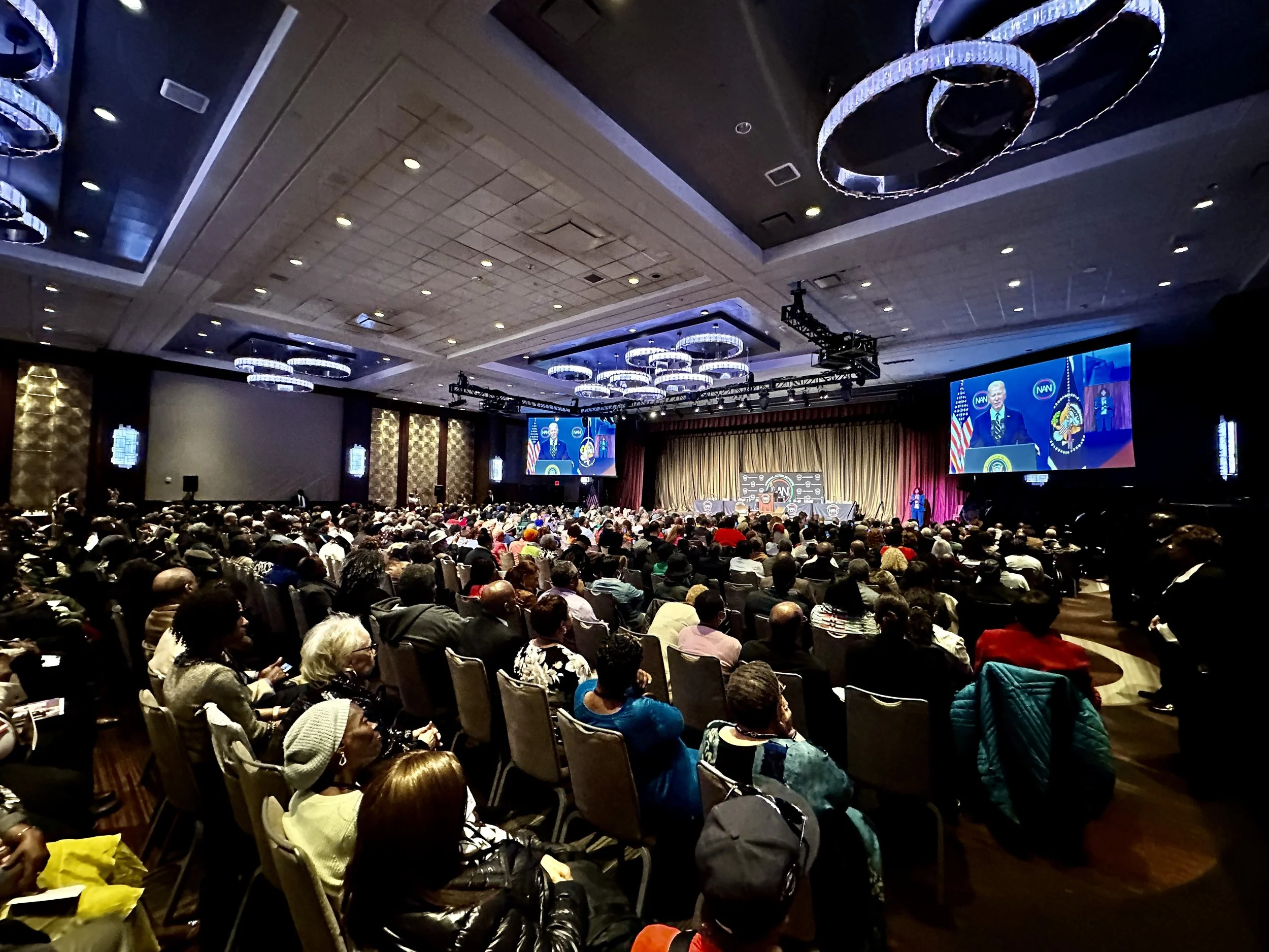 A large conference hall filled with a diverse audience attending a formal event, with a stage showing a speaker at a podium and a large screen displaying the speaker's image.