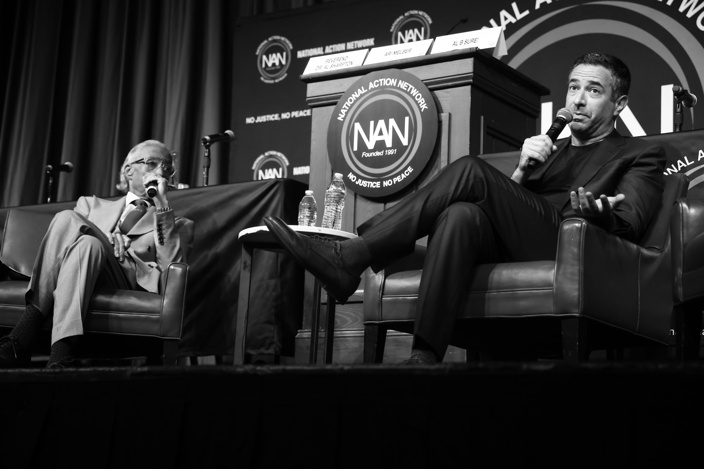 Two men are seated on stage at a discussion or interview event held by the National Action Network. One man on the left is an older individual with glasses and light-colored hair, while the man on the right is younger with dark hair, wearing a dark s