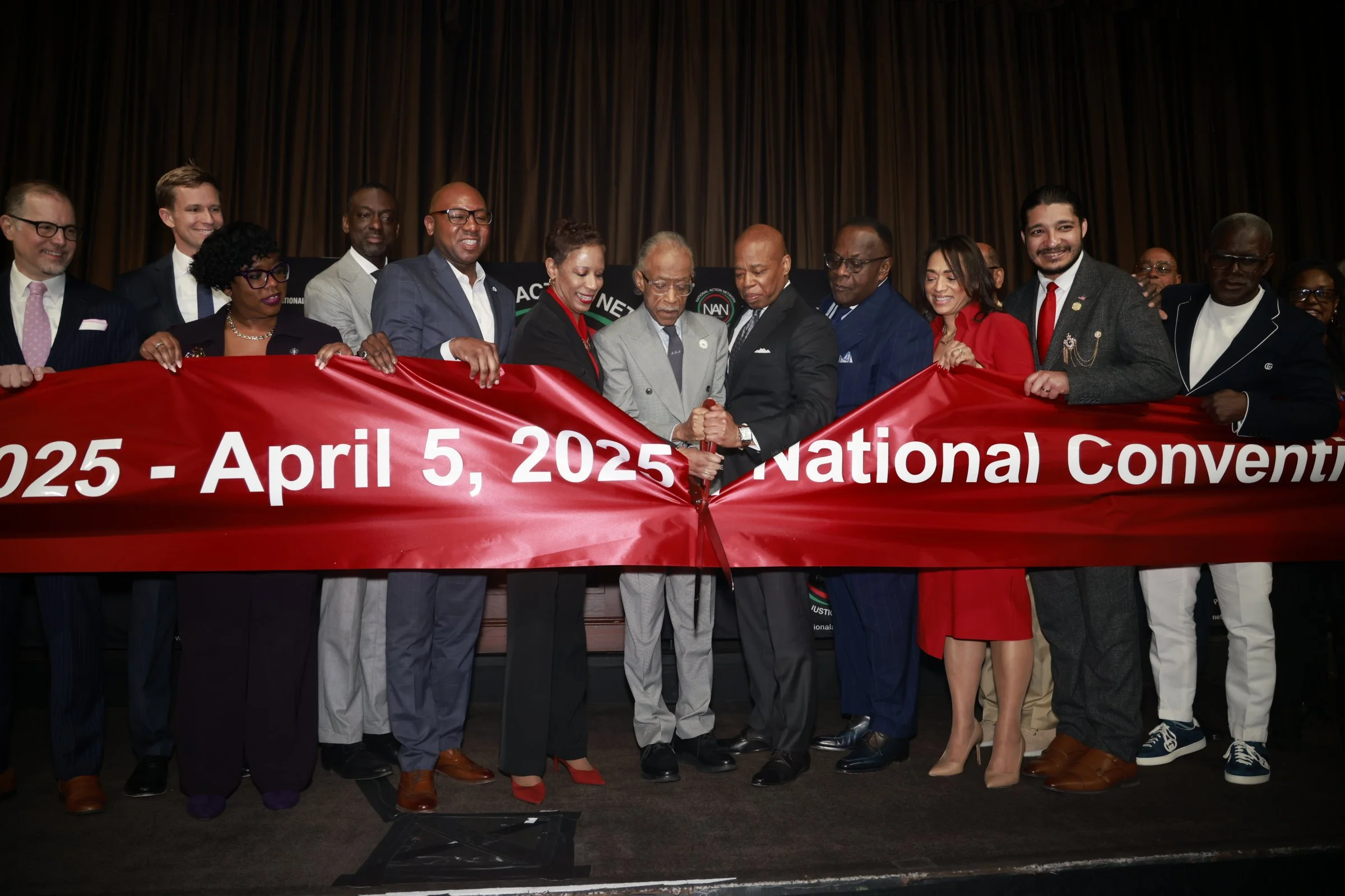 Group of people on stage participating in a ribbon-cutting ceremony for a 2025 National Convention, with a red ribbon and a large banner showing the event date.