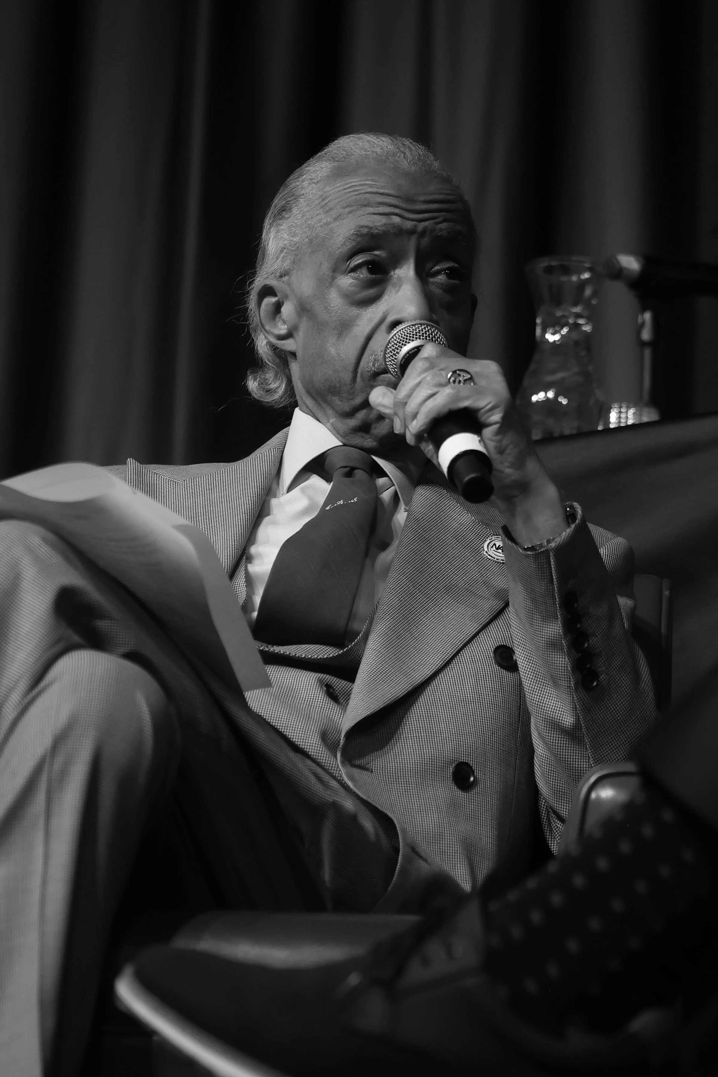 Black and white photo of an elderly man in a suit holding a microphone and reading from a piece of paper, with a dark curtain and glass pitcher in the background.