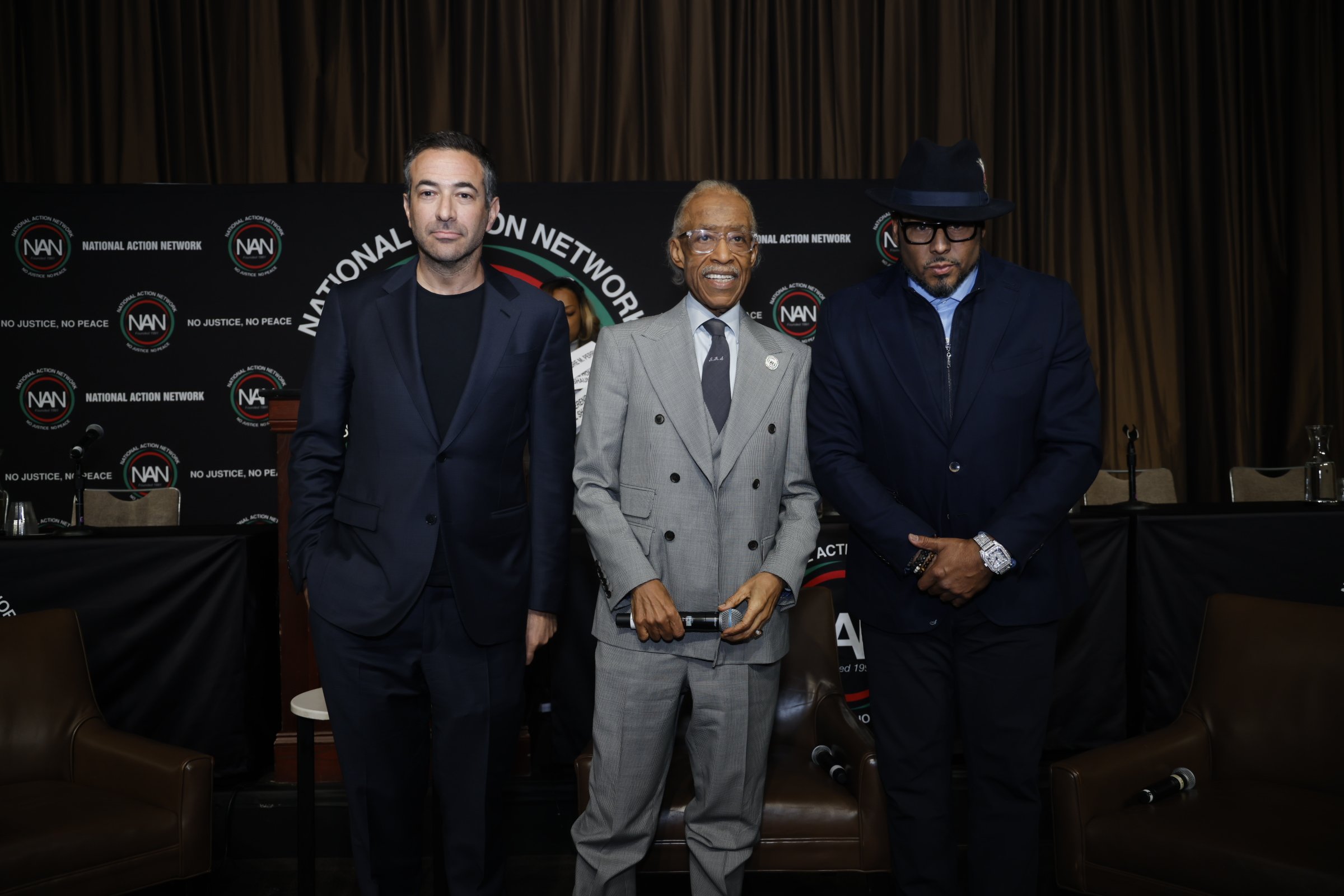 Three men stand together in front of a backdrop that says 'National Action Network' and 'No Justice, No Peace.' The man in the middle is holding a microphone and is wearing a light gray suit, the man on the left is in a dark suit with no tie, and the