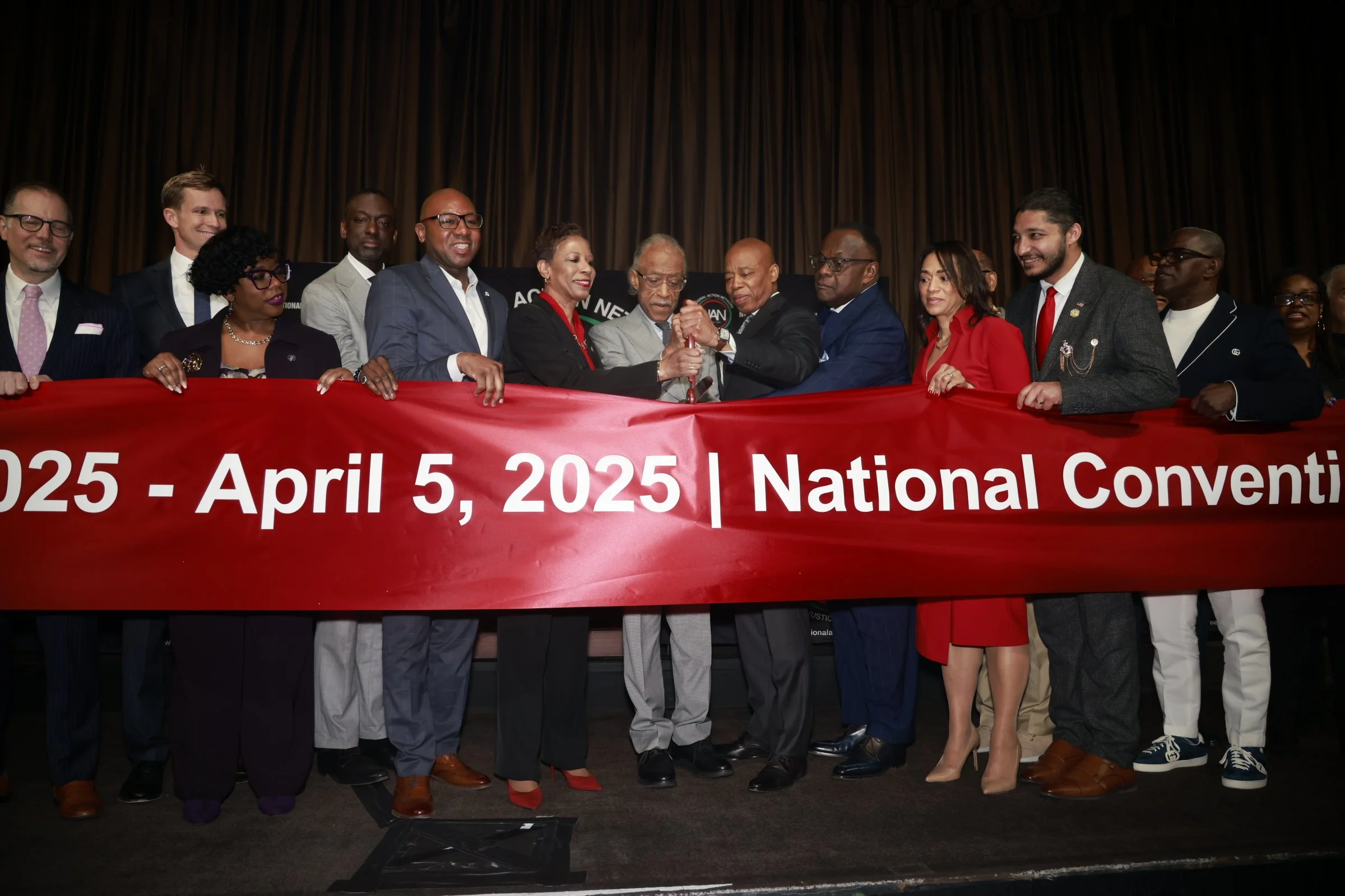 A diverse group of people holding a red banner with the text '2025 - April 5, 2025 | National Convention' during a formal event on a stage with a dark curtain backdrop.