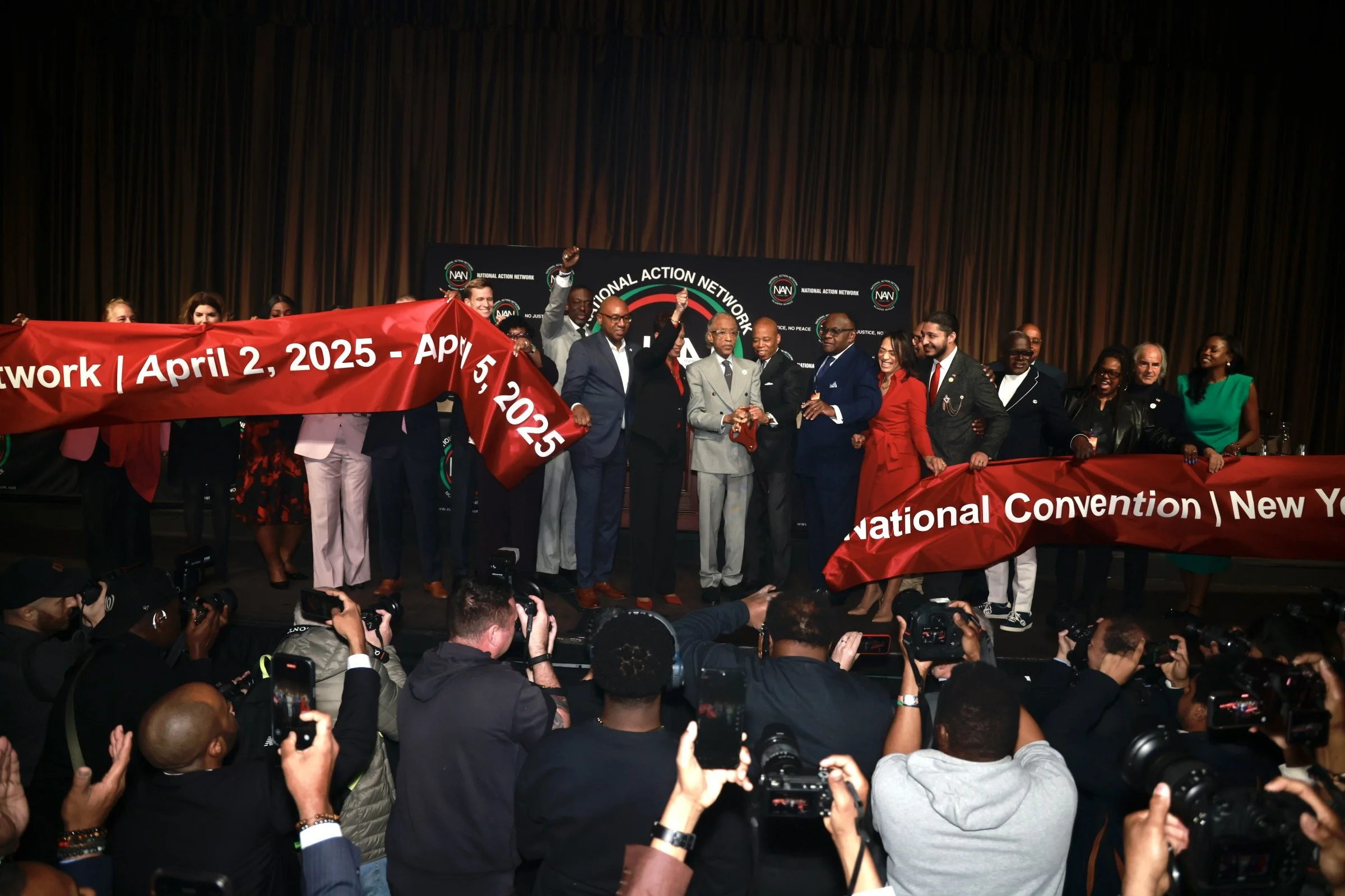 Group of people on stage holding a red banner at a conference, with photographers taking pictures in front.