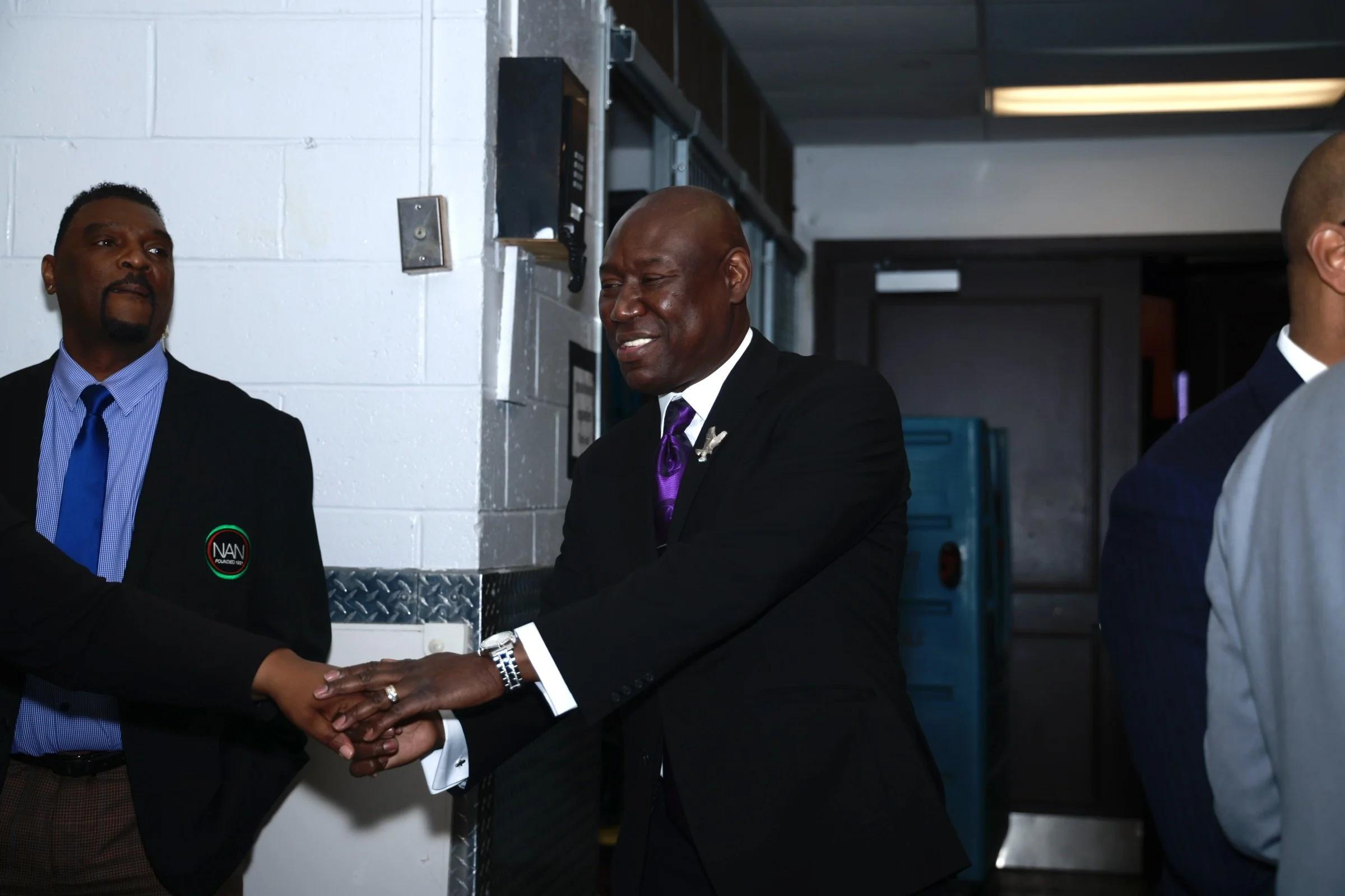 Two men in suits shaking hands in an indoor setting, with one man wearing a black suit and the other wearing a navy suit; the background includes white brick walls and blue lockers.