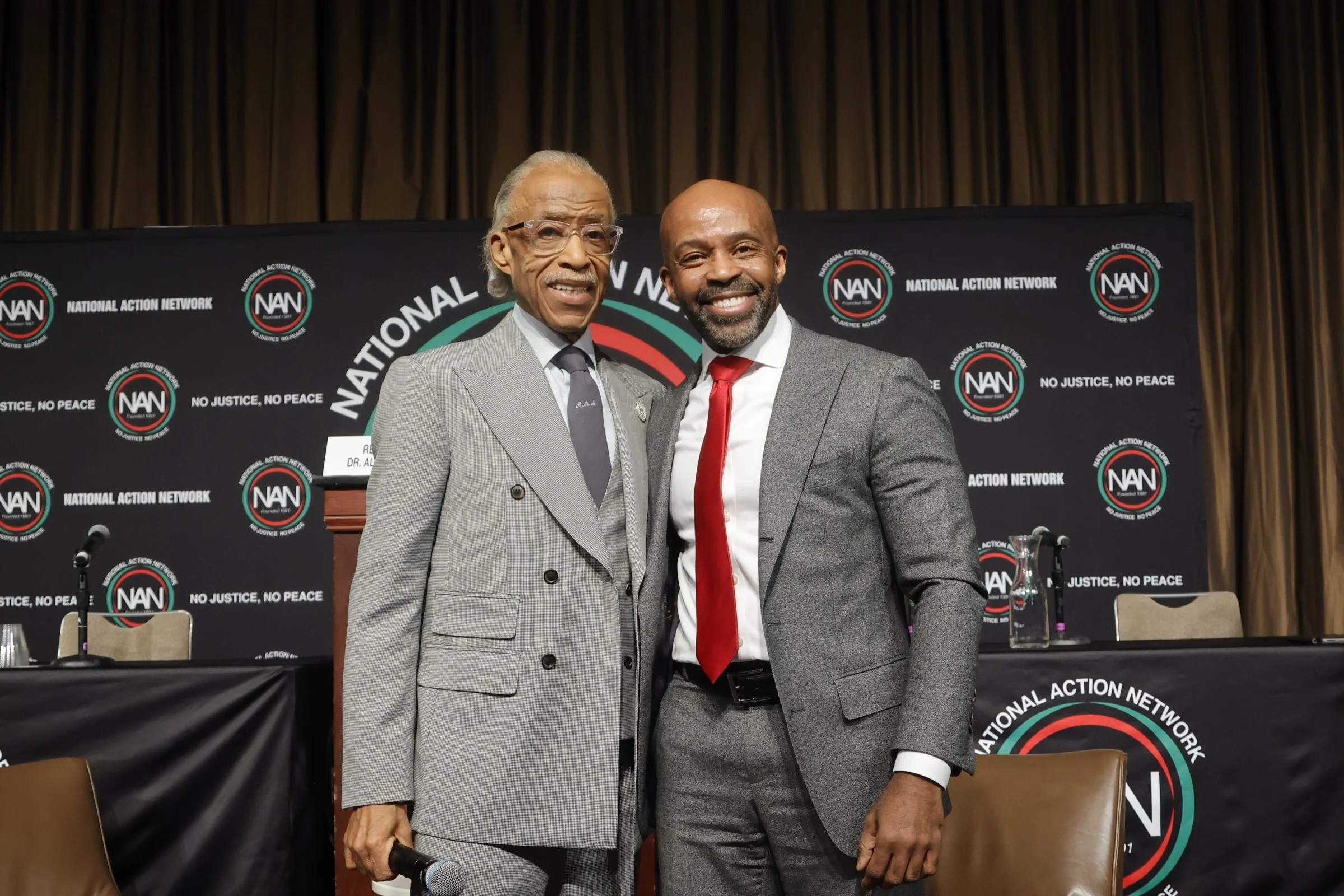 Two men in suits standing close together, smiling, in front of a banner with the logo of the National Action Network and the slogans 'No Justice, No Peace.'