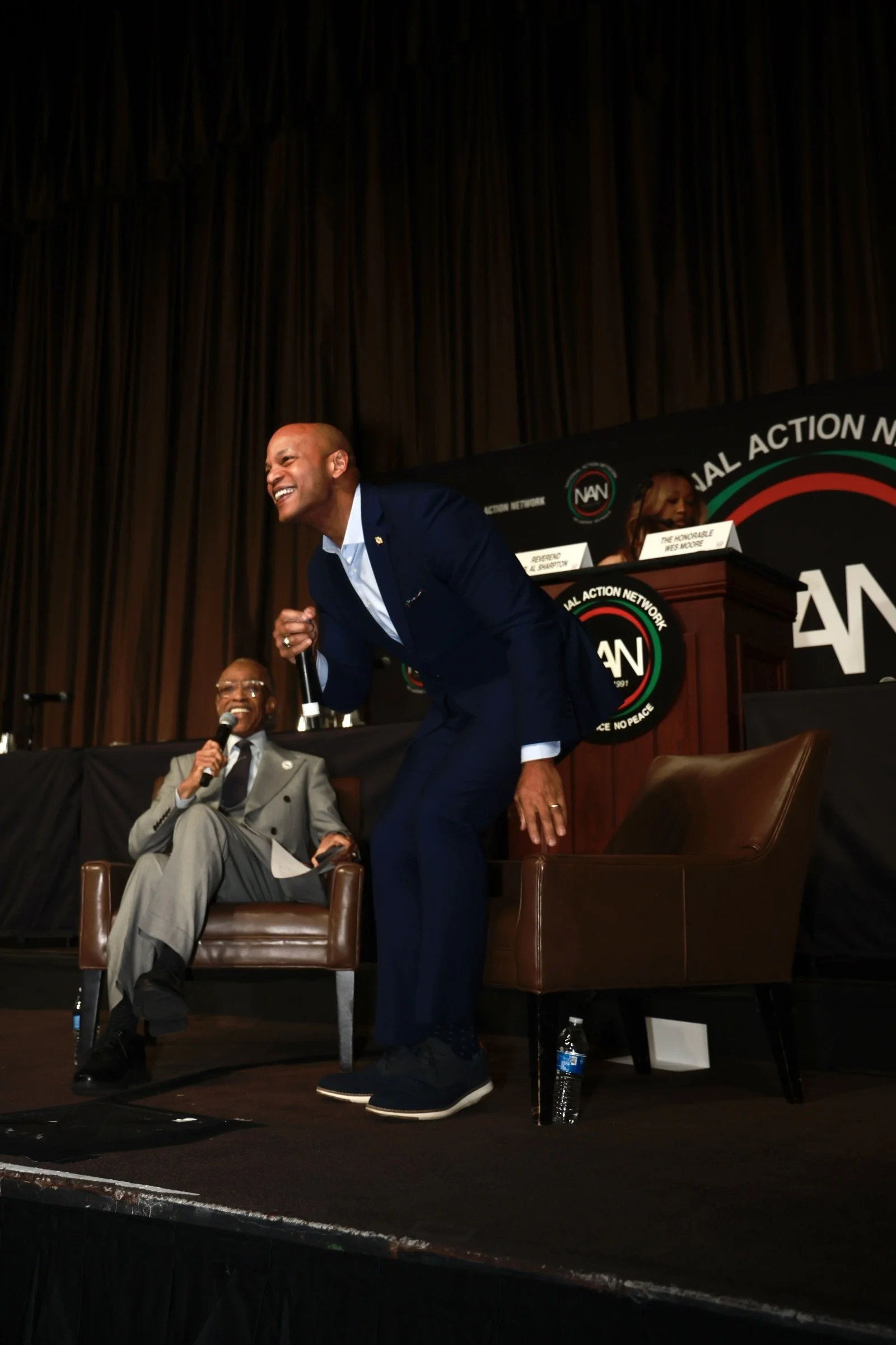 A man in a navy suit is standing on stage, leaning forward and laughing while holding a microphone. Another man in a gray suit is seated next to him, also laughing and holding a microphone. Behind them is a panel with individuals sitting at a long ta