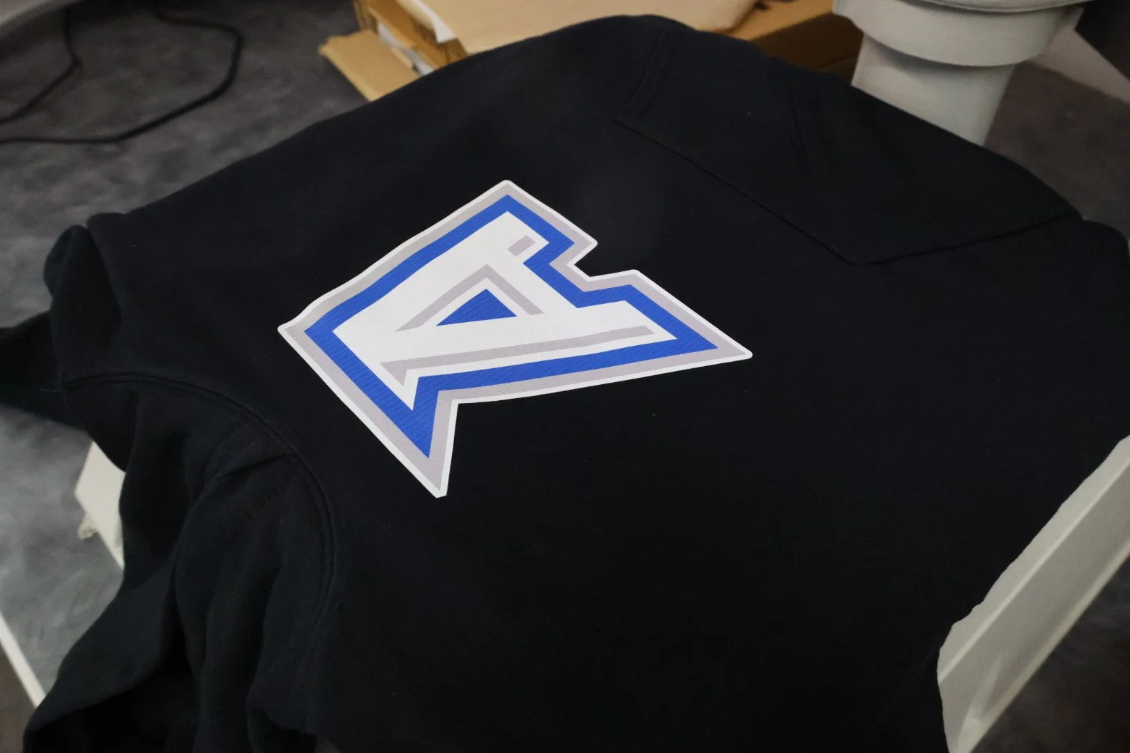 Black clothing item with a large white, blue, and gray logo on top, placed on a white table or surface.